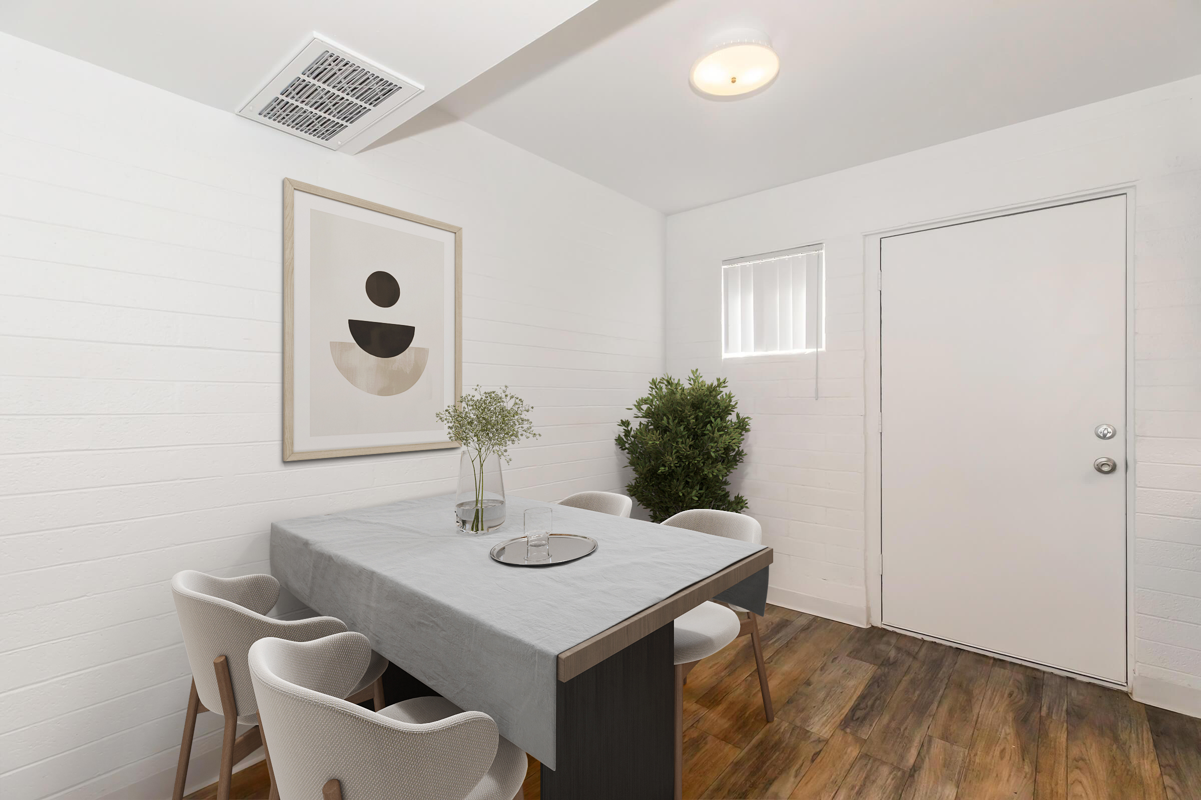 A modern, minimalist dining area featuring a rectangular wooden table with a light gray tablecloth. Around it are four white chairs. On the wall hangs an abstract art piece. A small plant is positioned in the corner, and a closed door is visible on one side, with natural light coming through a window.