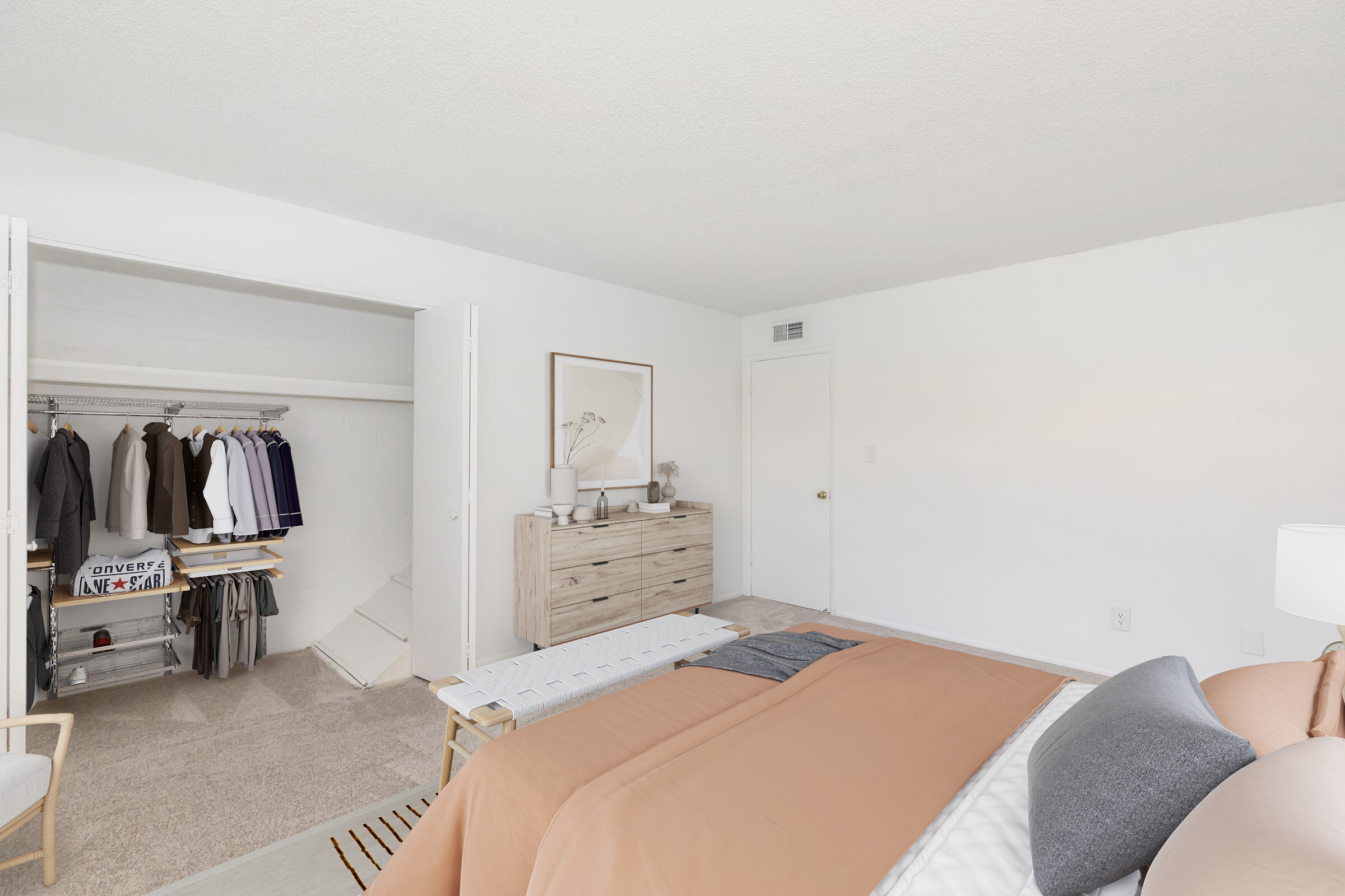 A bright, minimalist bedroom featuring a large bed with a brown and gray comforter. A wooden dresser is against one wall, and a closet with hanging clothes is visible on the left. The walls are painted white, and the carpeted floor adds warmth to the space. Natural light streams in, creating a cozy atmosphere.