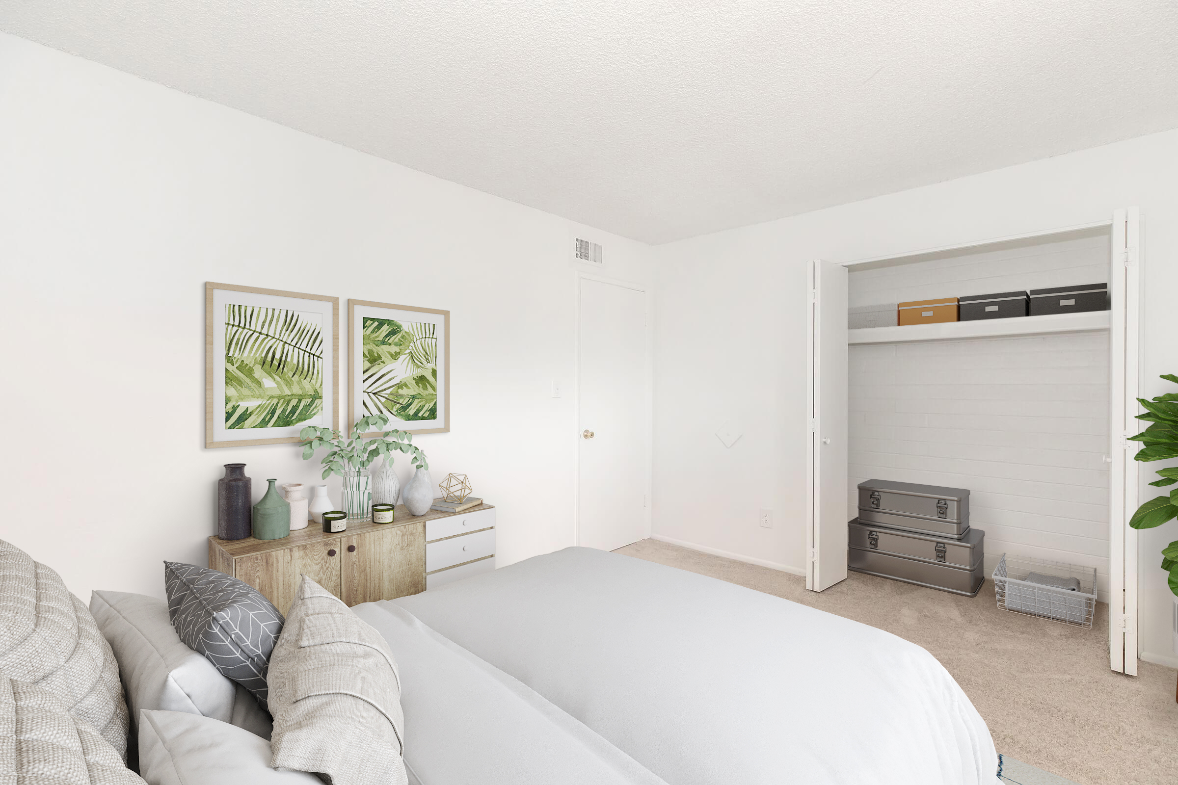 A bright and airy bedroom featuring a neatly made bed with gray linens, a bedside table decorated with plants and decorative containers, and two framed leafy prints on the wall. A walk-in closet is visible, with organized boxes and a potted plant adding a touch of greenery to the space.