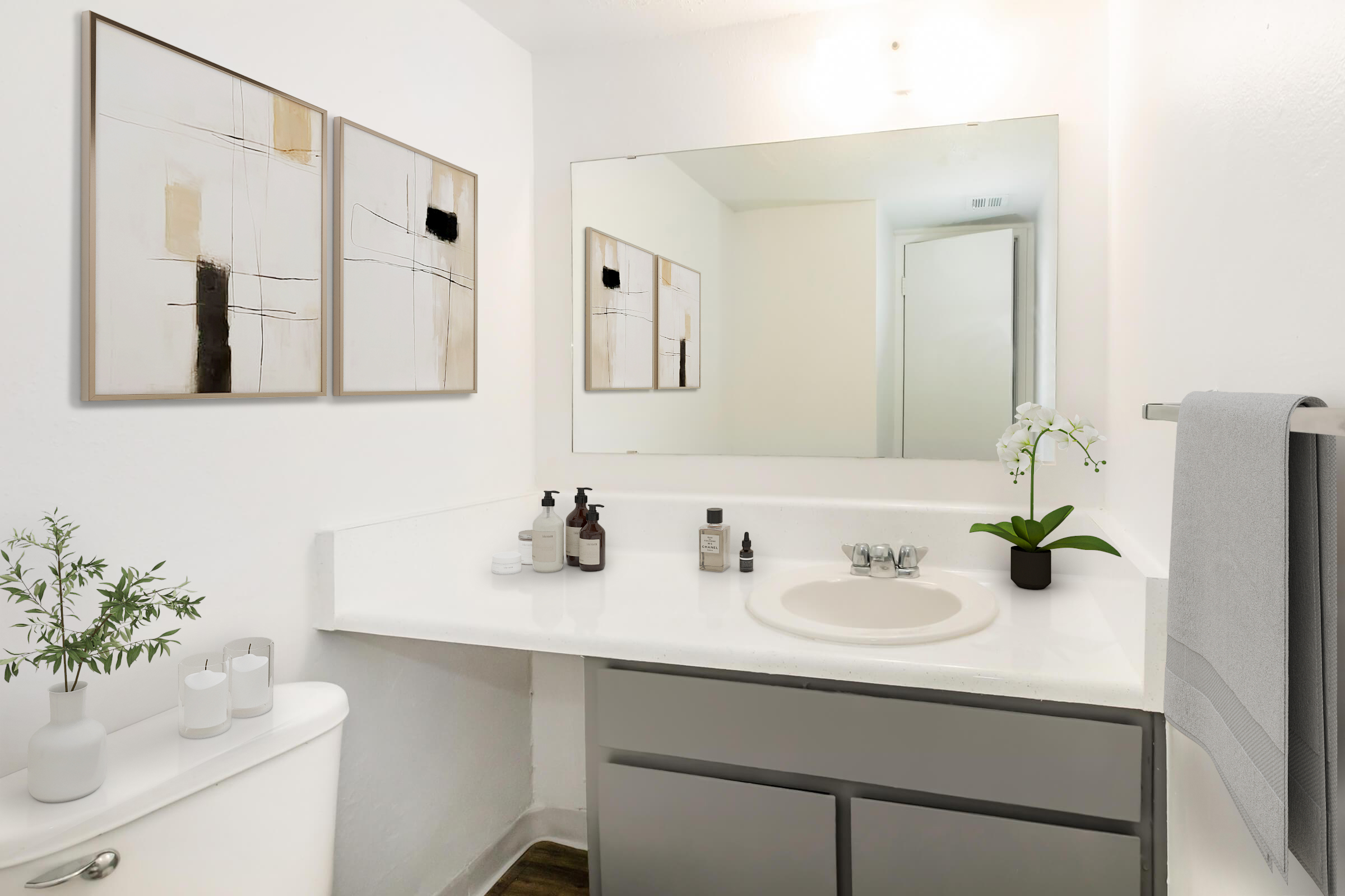 A modern bathroom featuring a clean white design. The space includes a white countertop with various skincare products, a small plant, and two framed abstract art pieces on the wall. A large mirror reflects the room's minimalistic decor, with a towel hanging on a rack and a toilet visible in the corner.