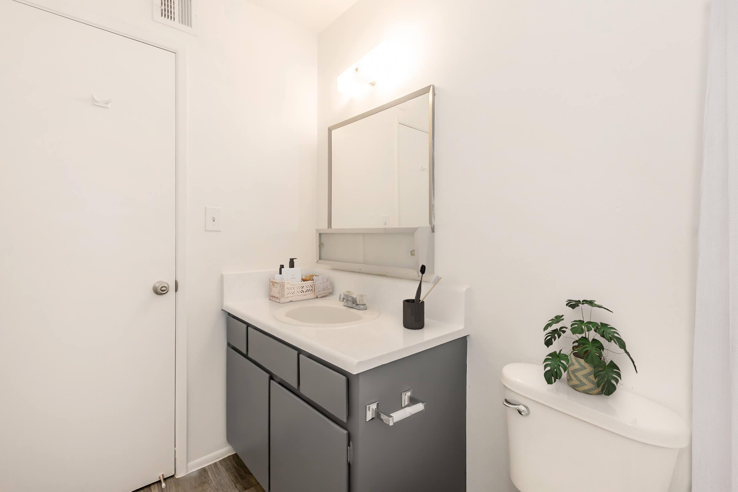 A modern bathroom featuring a gray vanity with a sink, a large mirror above, and a small plant on the countertop. The walls are white, and there is a white toilet visible. A door is seen to the left, and the flooring is light-colored wood. The overall aesthetic is clean and minimalistic.