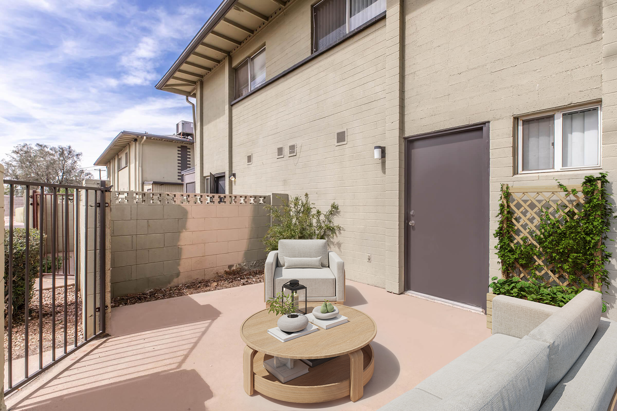 A cozy outdoor patio area featuring a light-colored sofa and armchair around a round coffee table. The space is enclosed by a fence and includes a wall adorned with climbing plants. A door leads into a building, and the sky is partly cloudy, providing a bright atmosphere.