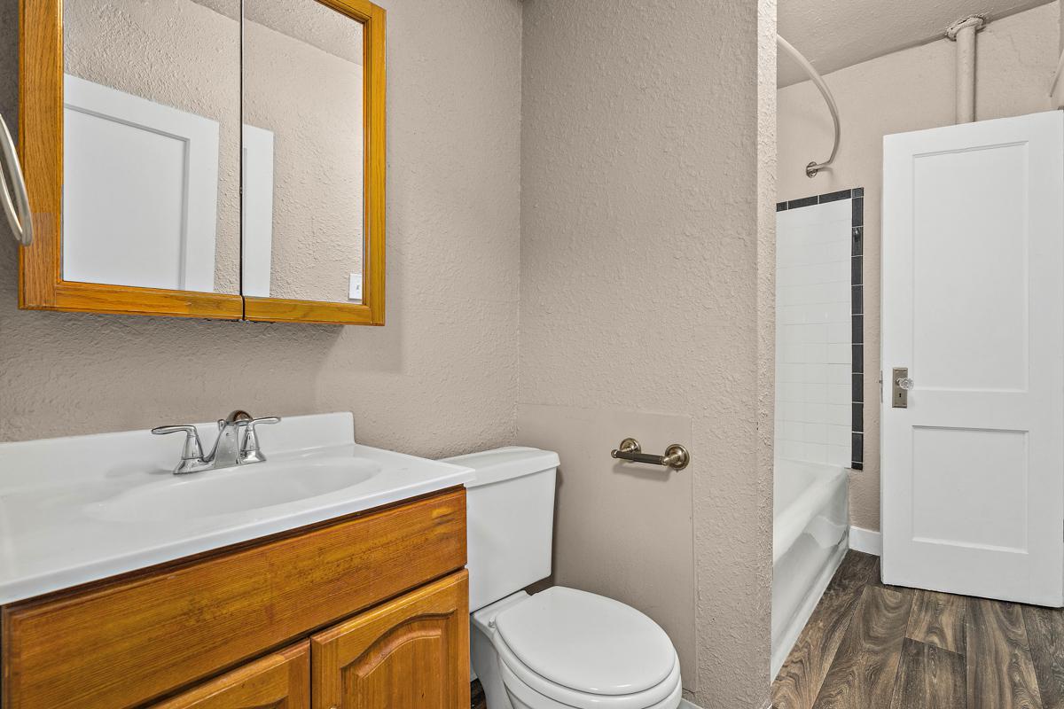 A small bathroom featuring a white vanity with a sink, a wooden cabinet, a toilet, and a bathtub with a shower. The walls are painted a neutral color, and there is a mirror above the sink. A door leads to another room, and the flooring is a dark wood laminate.