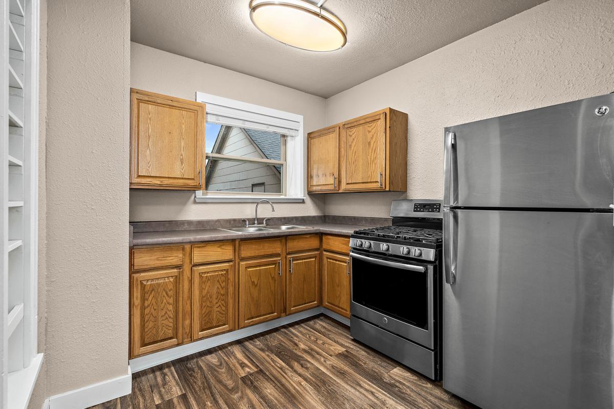 A small kitchen featuring light brown wooden cabinets, a stainless steel refrigerator, and a gas stove. There is a double sink beneath a window, with a neutral-colored wall and wooden flooring adding warmth to the space. The layout is compact and functional, ideal for a cozy cooking area.