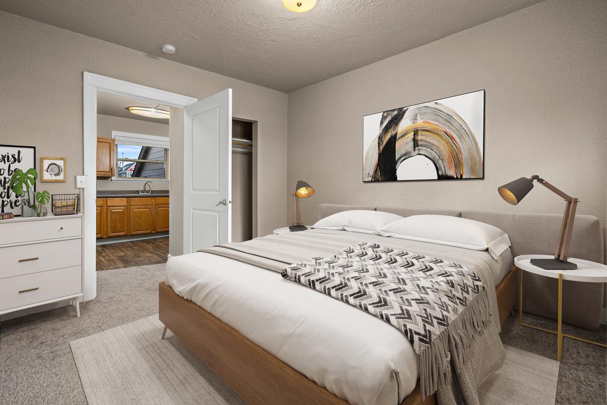 A cozy bedroom featuring a modern bed with neutral bedding and a patterned throw blanket. There's a bedside table with a lamp, a dresser with decorative items, and a door leading to a kitchen area in the background. The walls are painted in a soft color, and a piece of abstract art hangs above the bed.