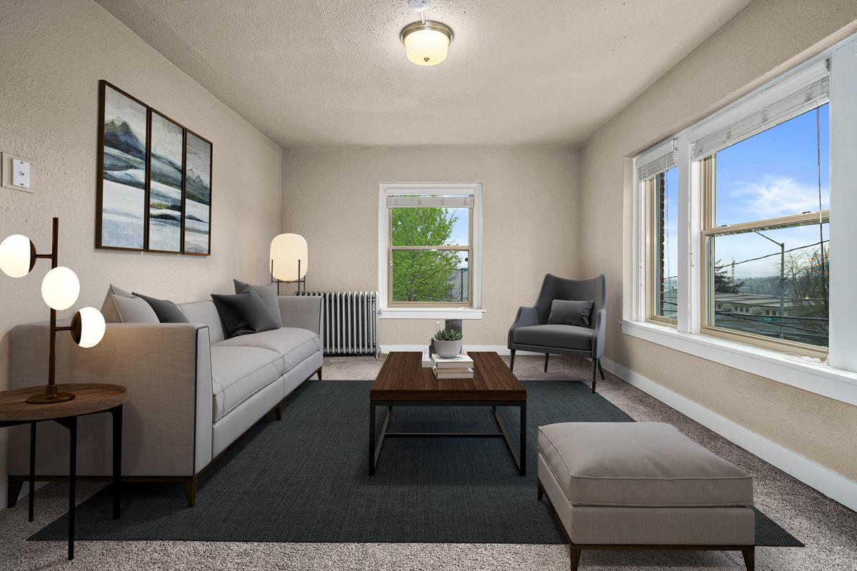 A cozy living room featuring a modern gray sofa, a wooden coffee table, and a plush ottoman. Large windows allow natural light to fill the space, showcasing views of greenery outside. The walls are painted a soft beige, and there are two abstract art pieces hanging above the sofa. A stylish floor lamp and a comfortable armchair complete the decor.