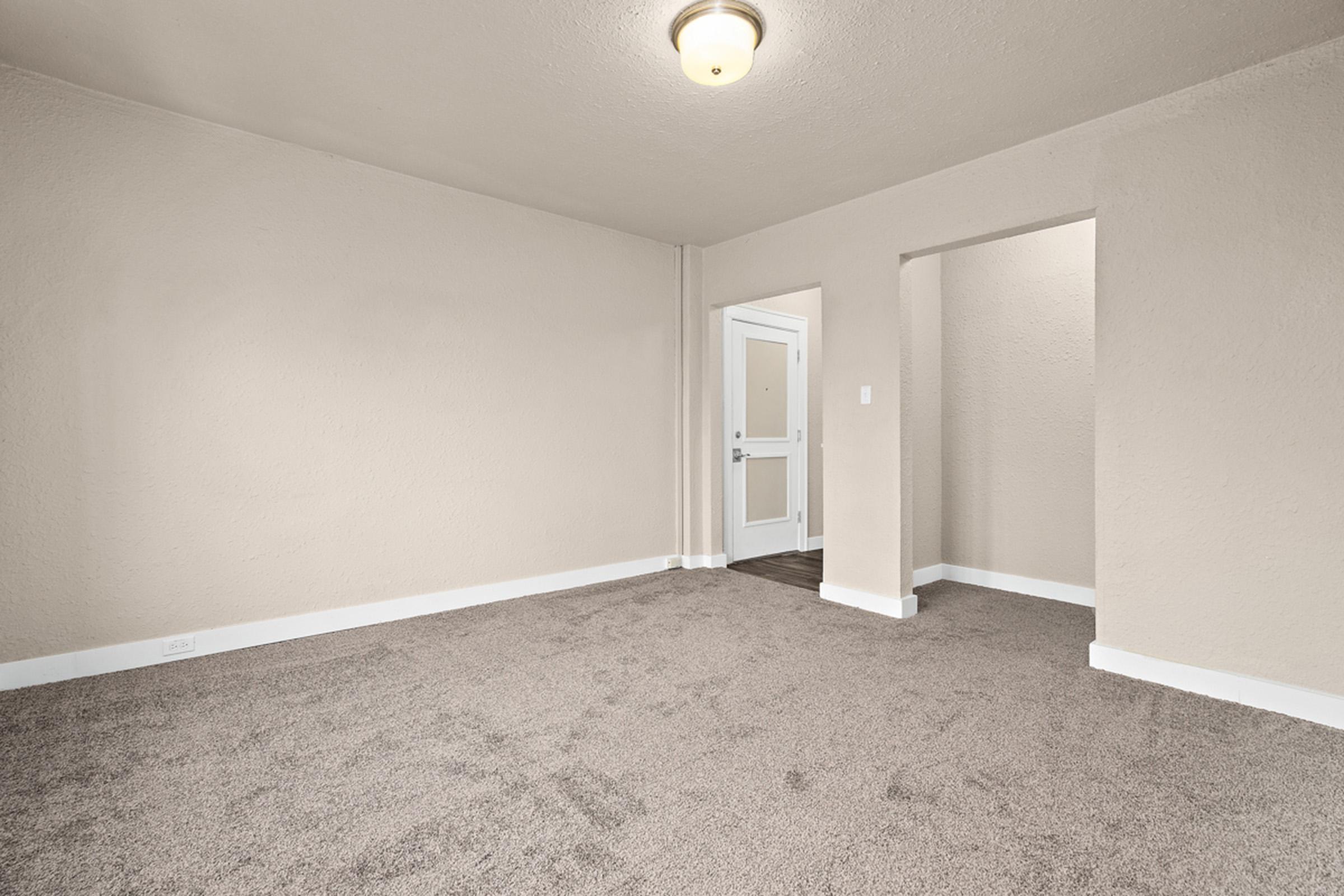 A spacious room with light beige walls and new gray carpeting. Natural light enters from a doorway on the left, leading to an entrance. The room has a simple ceiling light fixture and an alcove on the right.