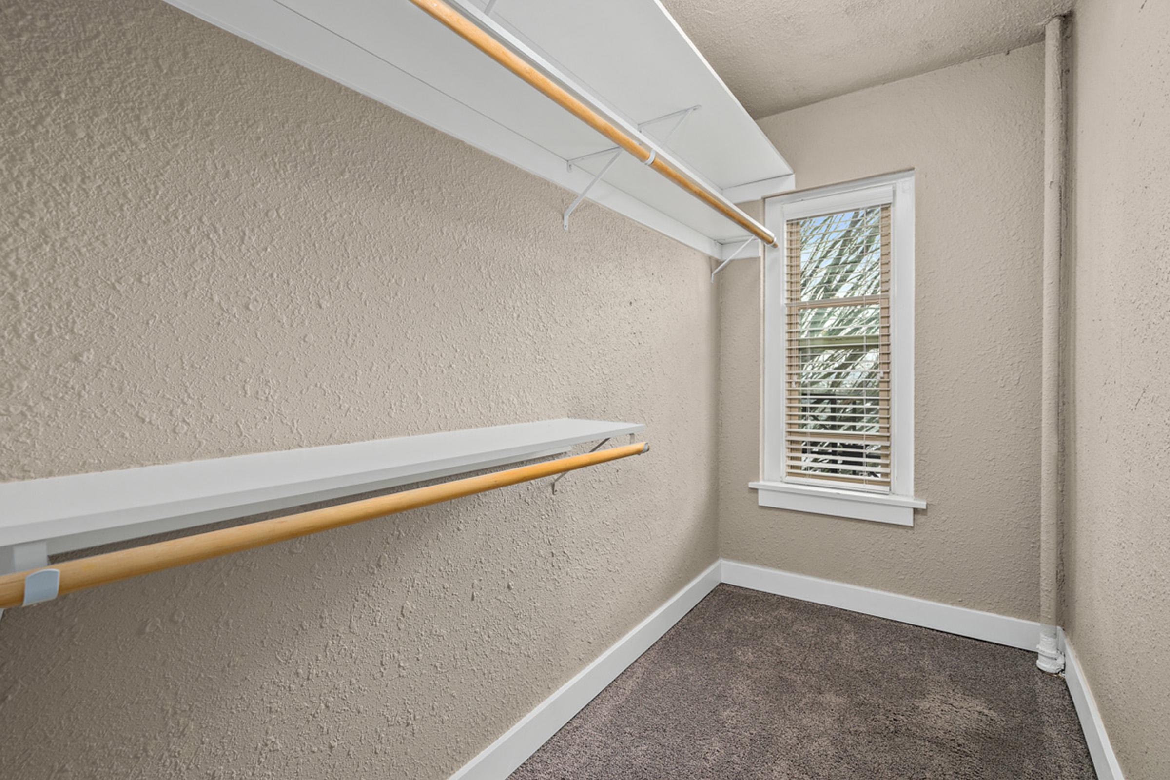 A small, empty closet featuring white shelves and a clothing rod against a light beige wall. Natural light filters through a partially open window with white blinds. The floor is carpeted in a neutral color, creating a bright and clean storage space.
