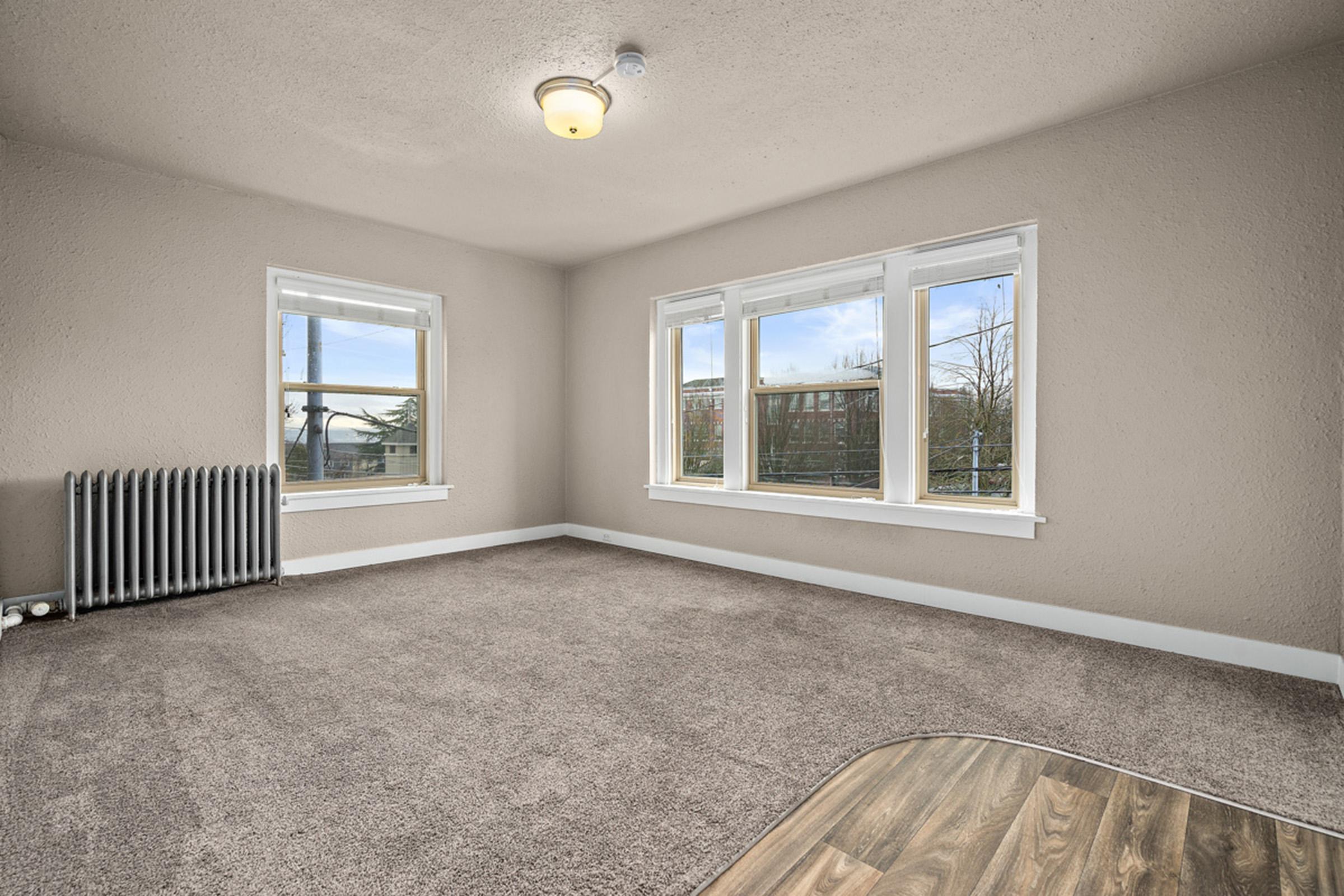 A spacious, empty room with light gray walls and large windows allowing natural light. There is a light fixture on the ceiling, a radiator along one wall, and carpeted flooring. The room features a wooden edge on the floor, creating a contrast with the carpet.