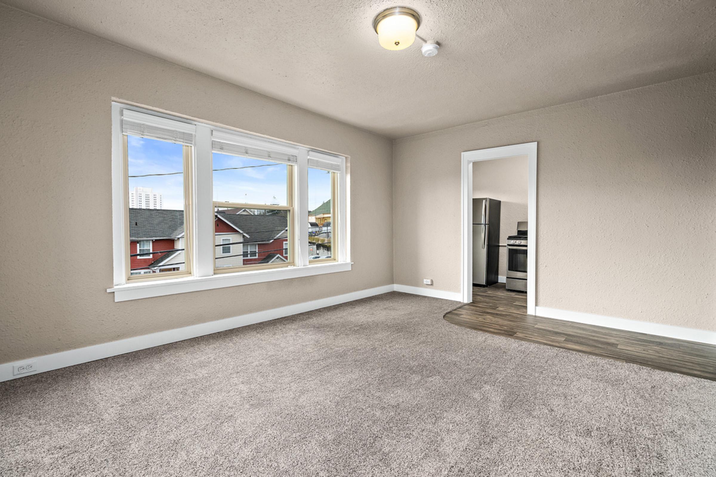 A bright, empty room with light gray walls and a plush carpet. Large windows allow natural light to fill the space, showcasing a view of rooftops outside. A doorway leads to a kitchen area, visible in the background, featuring an appliance. The ceiling has a simple light fixture.