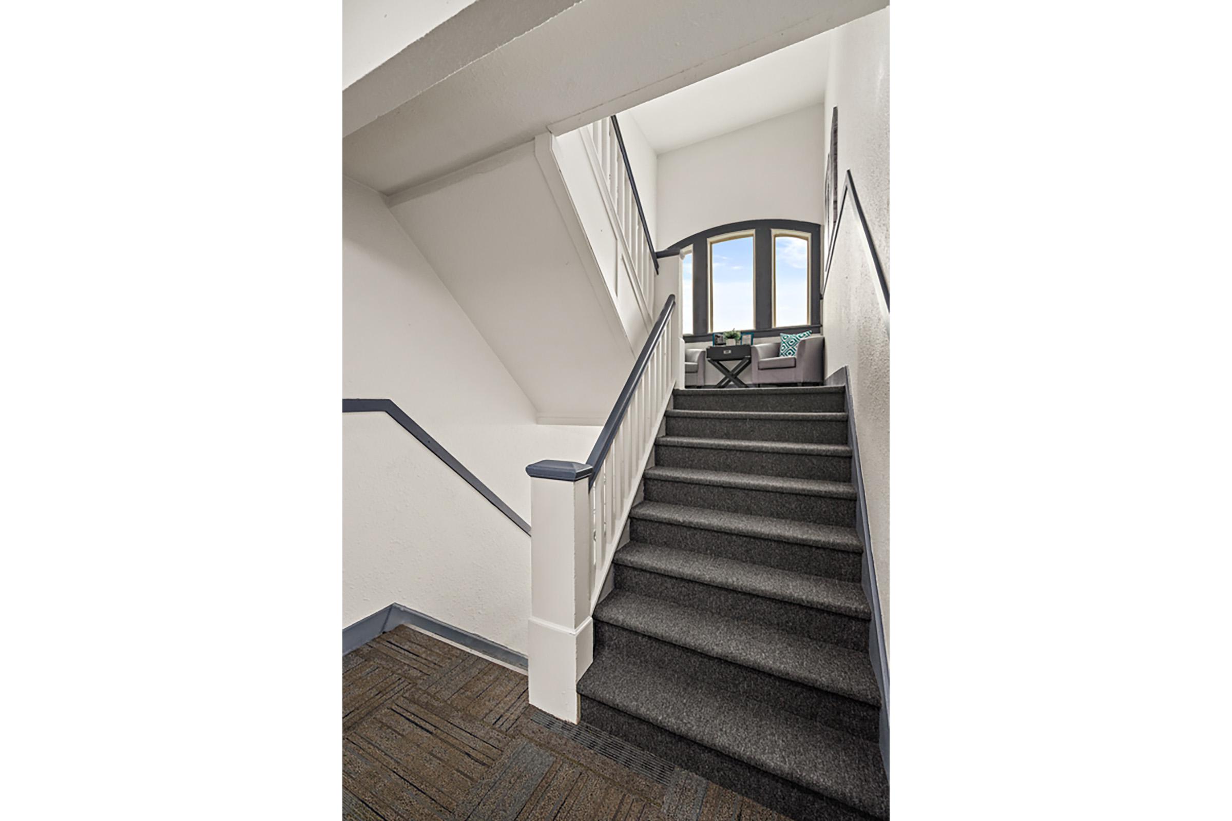 Interior view of a staircase leading upwards. The stairway features gray carpeting and white walls with blue trim. Natural light streams in from a large window at the top, illuminating a seating area with a small table and chairs. The overall design is modern and inviting.