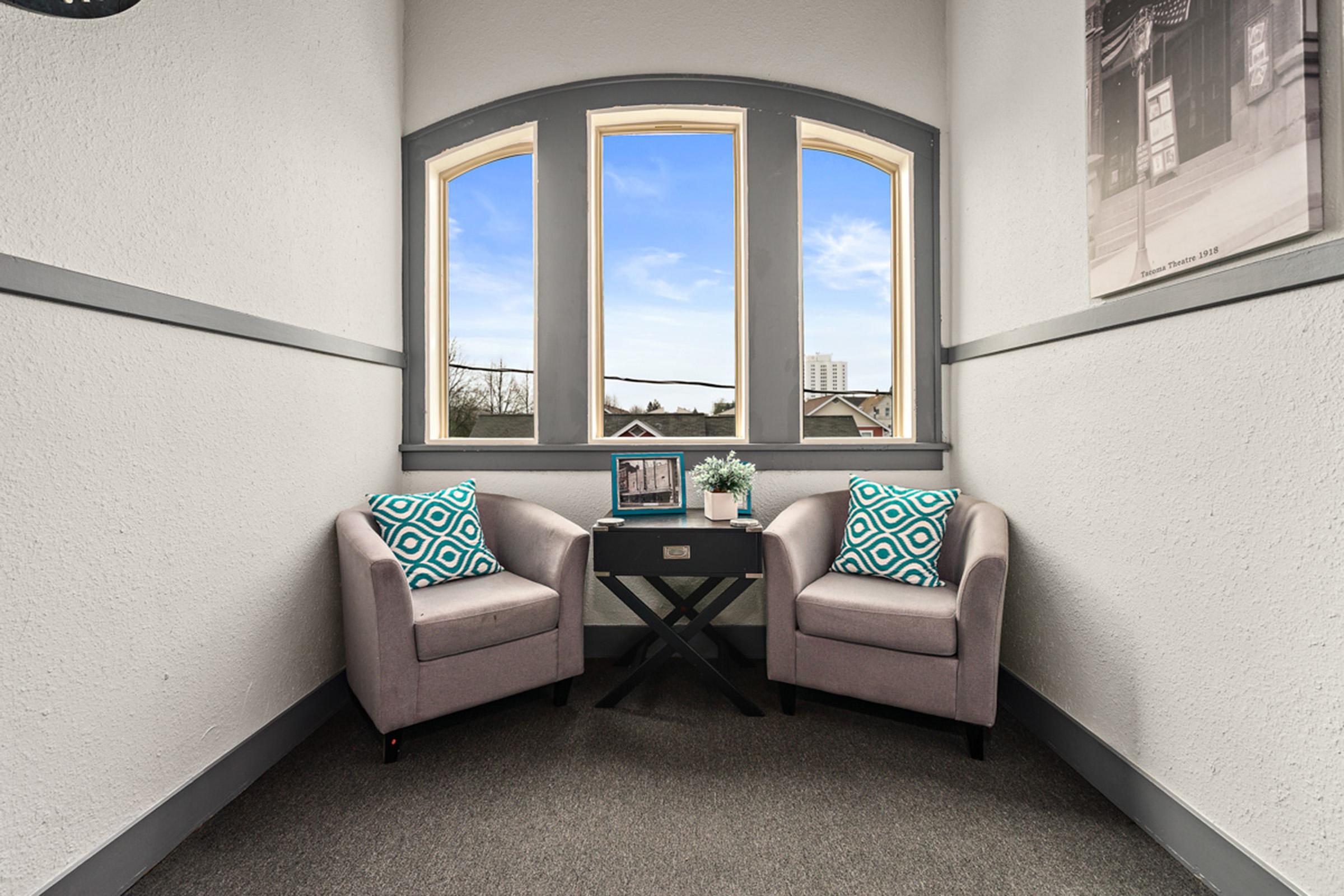 A cozy corner featuring two light gray armchairs with patterned cushions, positioned near large windows that provide a view of the outdoors. A small black side table sits between the chairs, adorned with a decorative plant and a framed photo, against a backdrop of textured walls and gray trim.