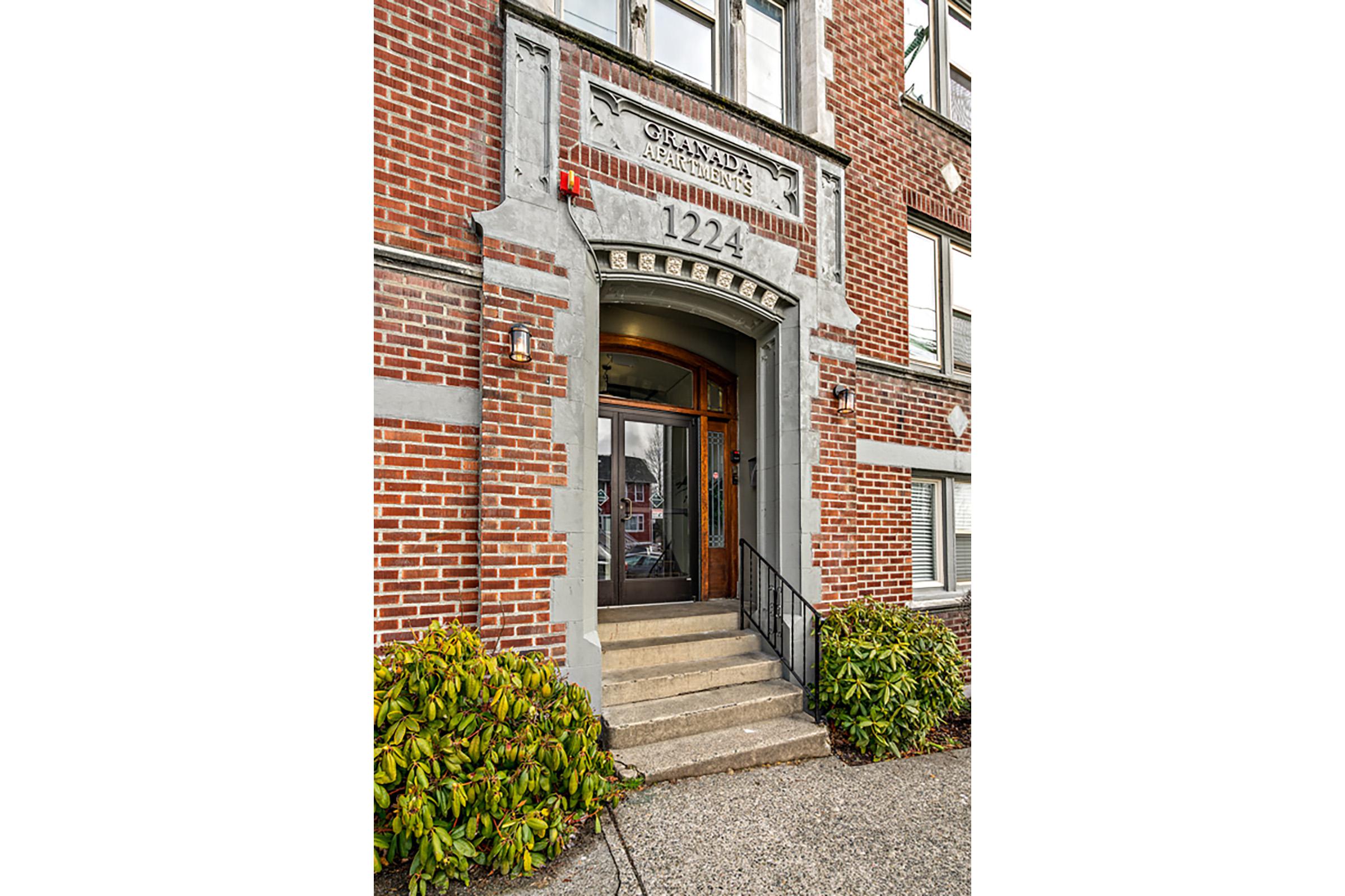 Entrance of a vintage brick apartment building with a decorative stone archway. The building number "1224" is prominently displayed above the wooden double doors. A set of steps leads up to the entrance, flanked by leafy green shrubs on either side. The overall architecture reflects a classic early 20th-century style.