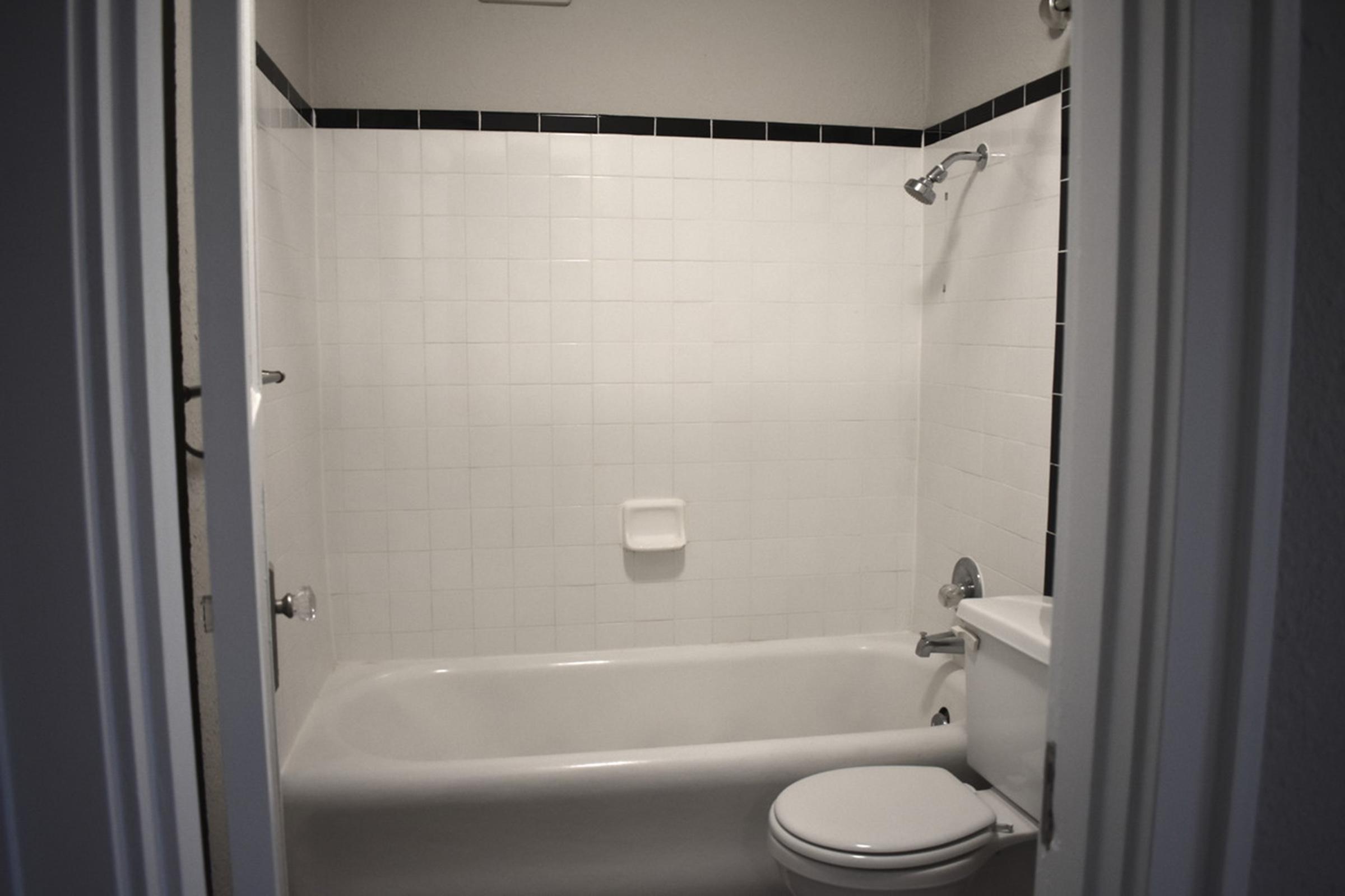 A clean, minimalistic bathroom with white tiled walls, a bathtub, a showerhead on the wall, and a toilet. The space has black accent tiles along the top, creating a modern look. The doorframe is visible, suggesting the entrance to the bathroom.