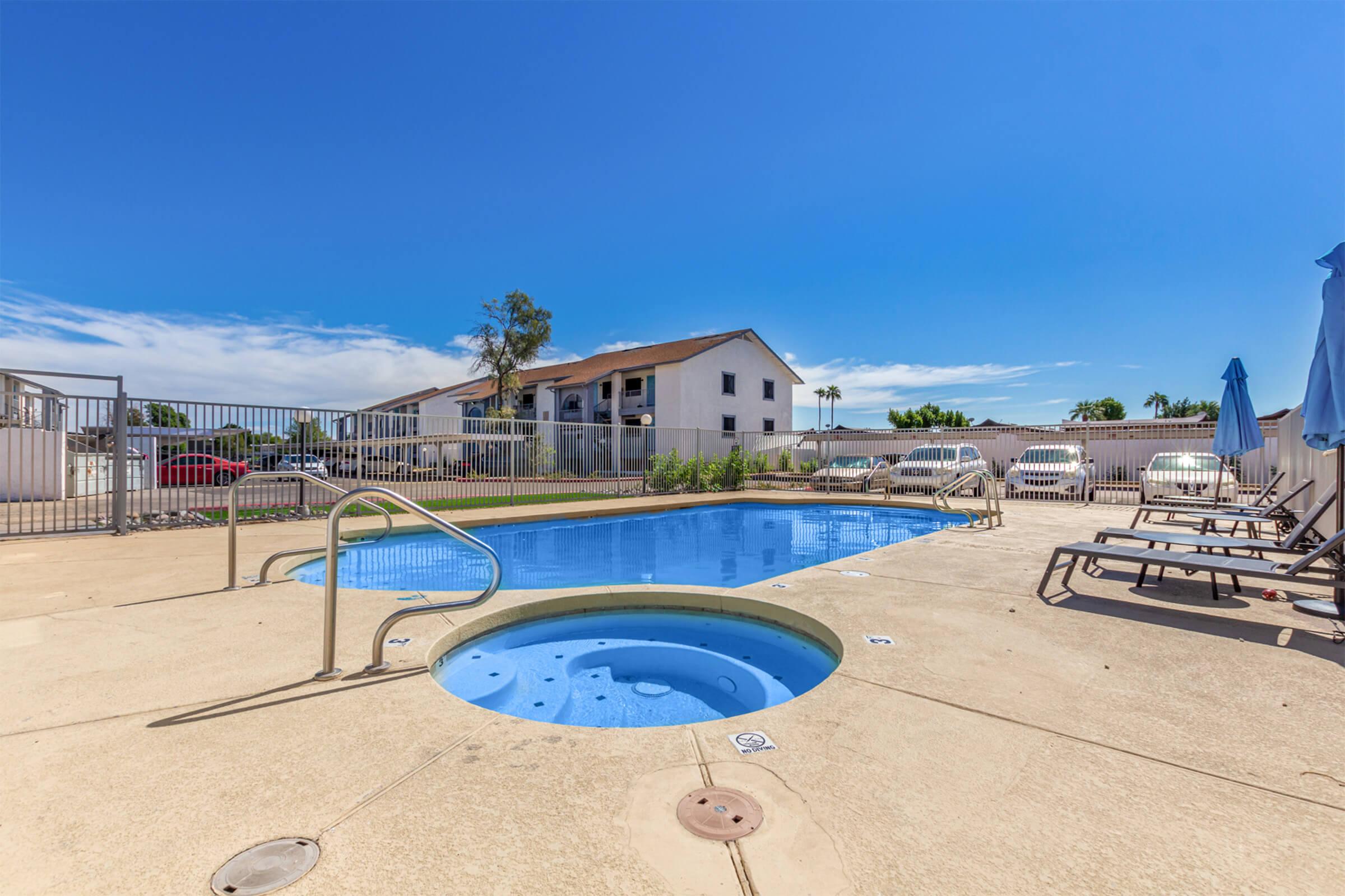 A clear blue swimming pool and a small hot tub, surrounded by a pool deck with lounge chairs. In the background, there's a white building with multiple windows, under a bright blue sky. A few cars are parked nearby, and the setting appears sunny and inviting.