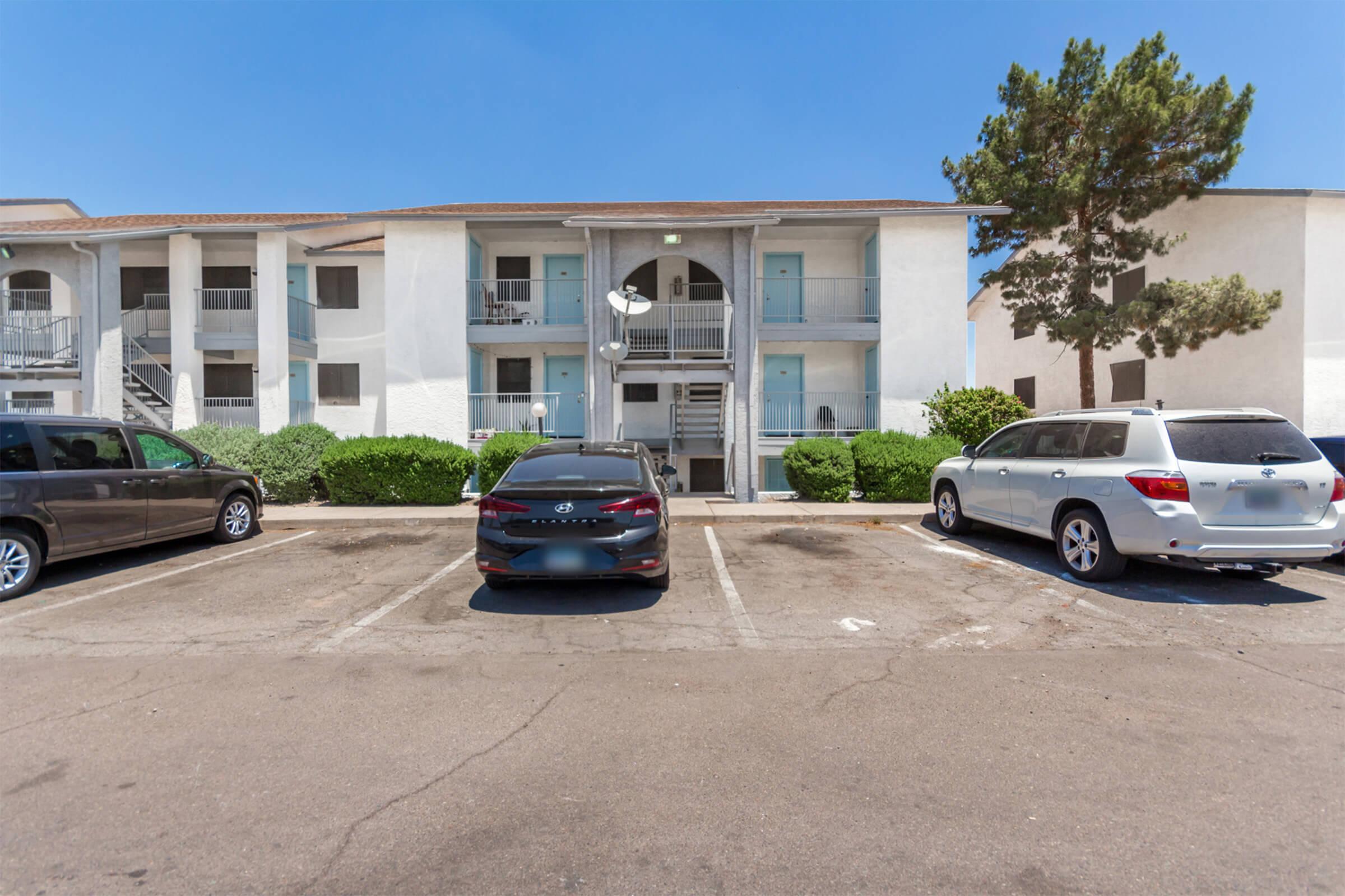 Two-story apartment building with a light-colored exterior, featuring balconies. A small parking lot in front has several cars parked, including a dark sedan and a silver SUV. Lush green bushes line the building, and a tree is visible on the right side of the image. The sky is clear and blue.