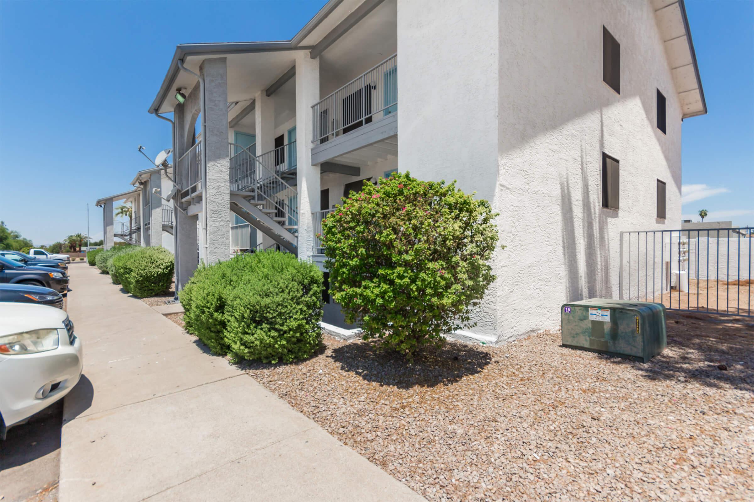 A view of a residential apartment building featuring a light-colored exterior, balconies, and a pathway lined with bushes. Parking spaces are visible on the left, and there is a utility box near the building. The sky is clear and sunny.