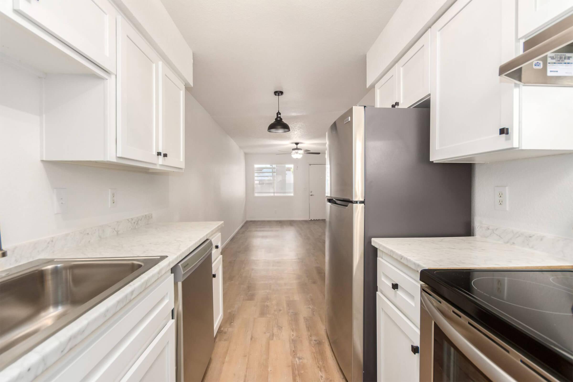 A modern kitchen featuring stainless steel appliances, including a fridge, oven, and dishwasher. White cabinetry and countertops complement the light wood flooring, creating a bright and spacious atmosphere. The room is well-lit with overhead lighting, and a doorway leads to an adjoining area.