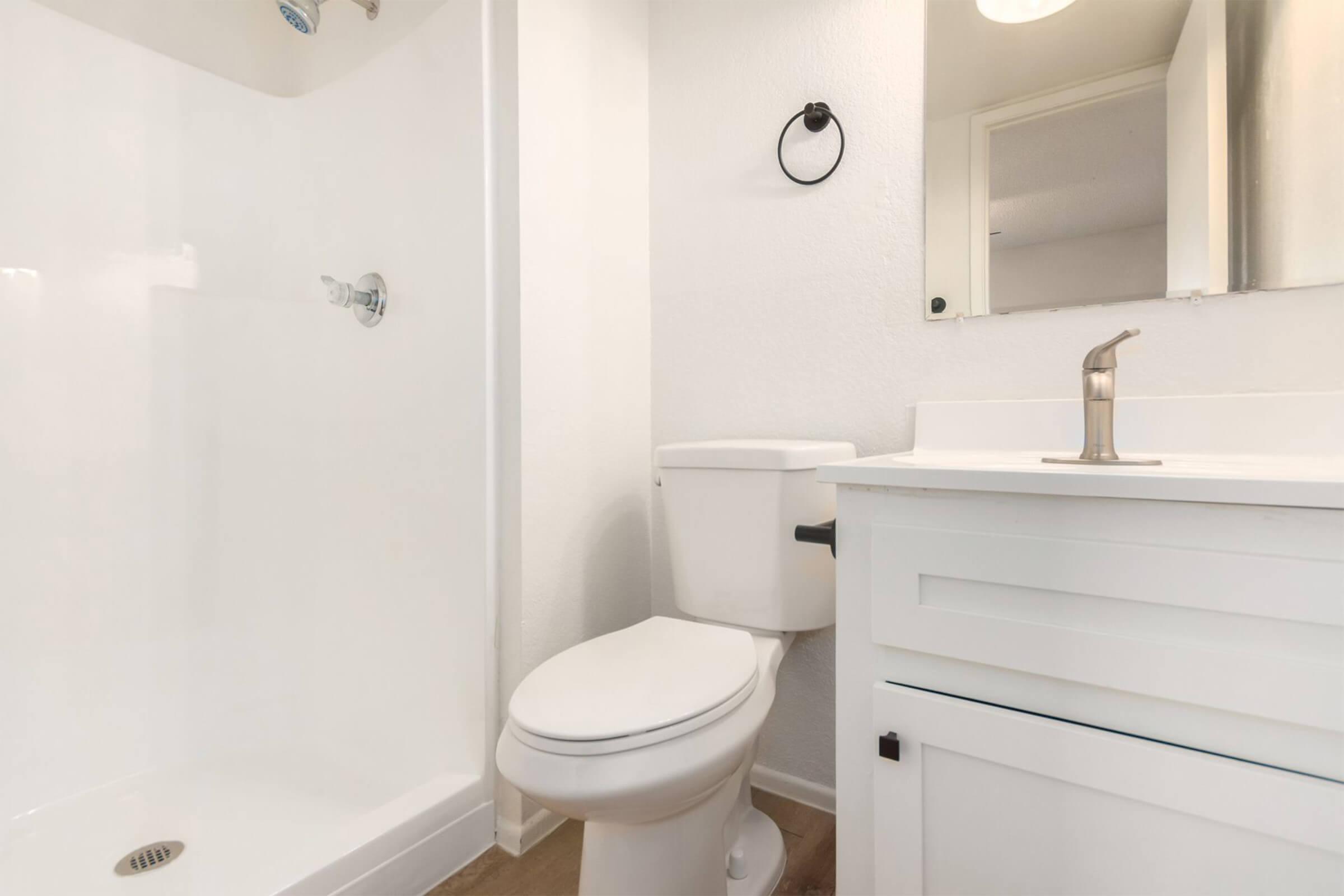 A clean, modern bathroom featuring a standalone shower, white toilet, and a sink with a mirror above. The walls and fixtures are bright, creating a spacious and inviting atmosphere. A small round towel holder is mounted on the wall. The floor is light-colored, enhancing the overall brightness of the space.
