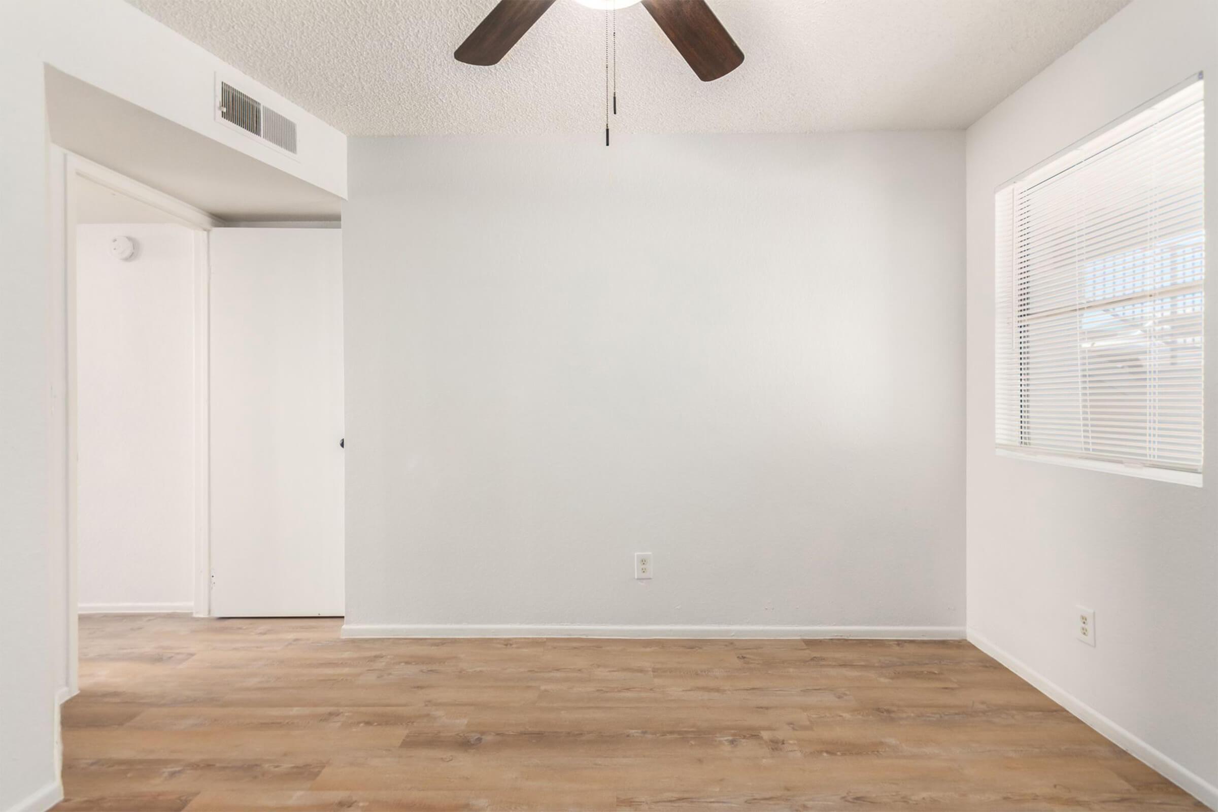 A bright, empty room with light wooden flooring and a white wall. A ceiling fan is visible, and there's a window letting in natural light. A closed door can be seen on the left, leading to another space, while the overall ambiance is clean and minimalistic.
