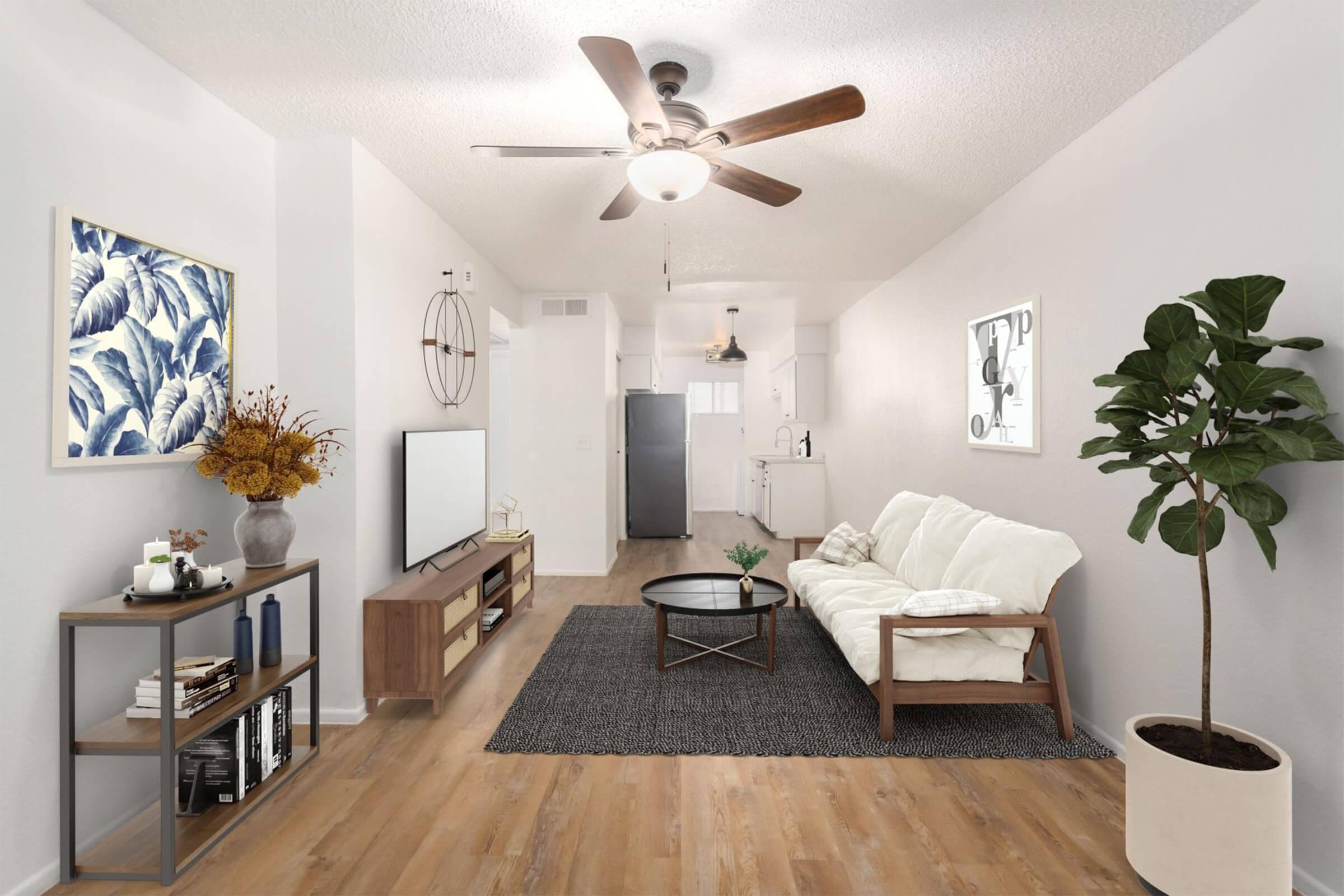 A modern living room featuring a white couch, a black coffee table, and a decorative rug. A wall-mounted television is positioned on a wooden console. Decorative plants and artwork enhance the space, with kitchen appliances visible in the background. Natural light fills the room, creating an inviting atmosphere.