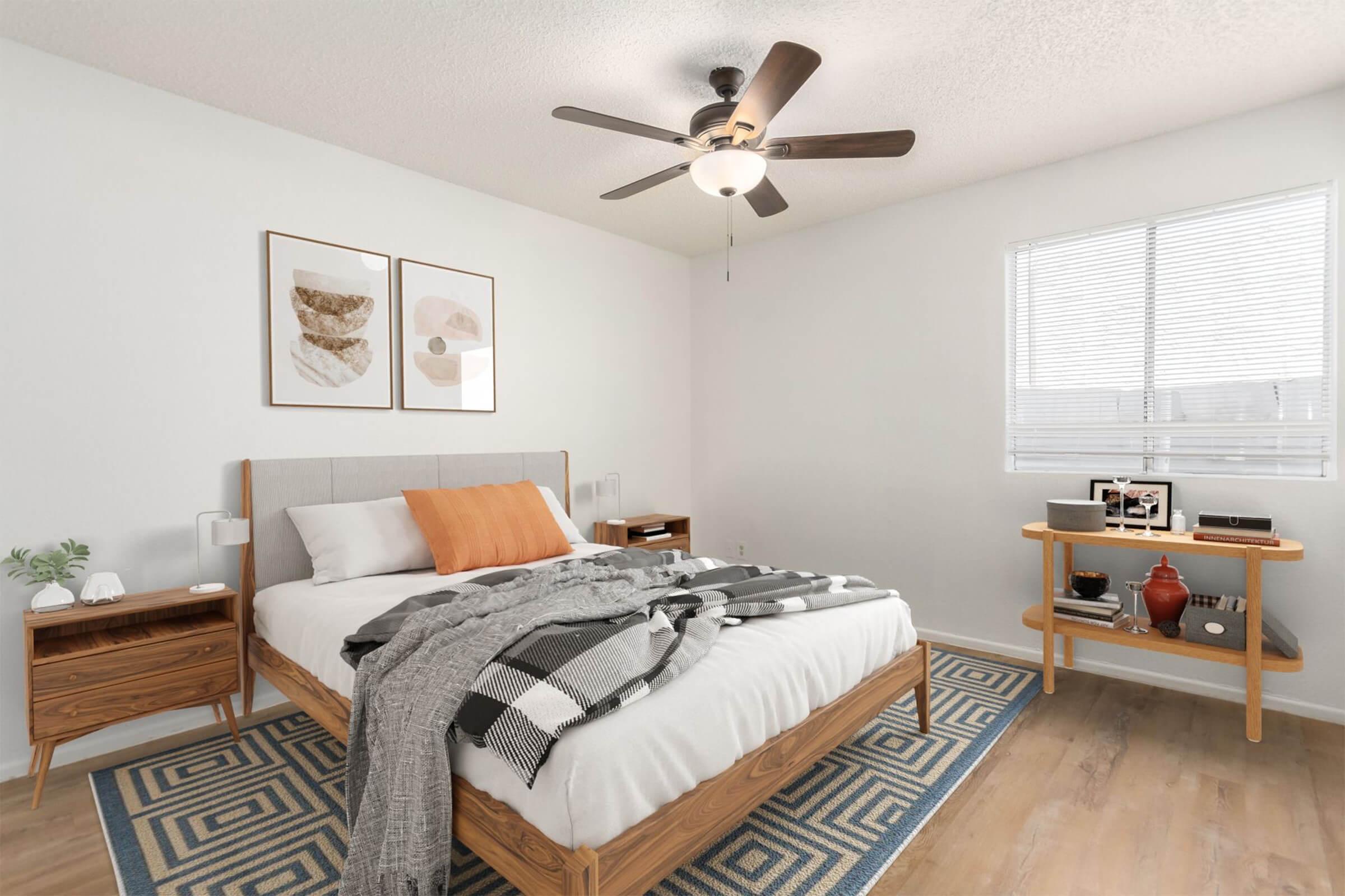 A cozy bedroom featuring a wooden bed with white bedding and a plaid blanket. There's a fan on the ceiling, framed artwork on the walls, and a small wooden side table with decor. A large window provides natural light, and the floor has a patterned area rug.