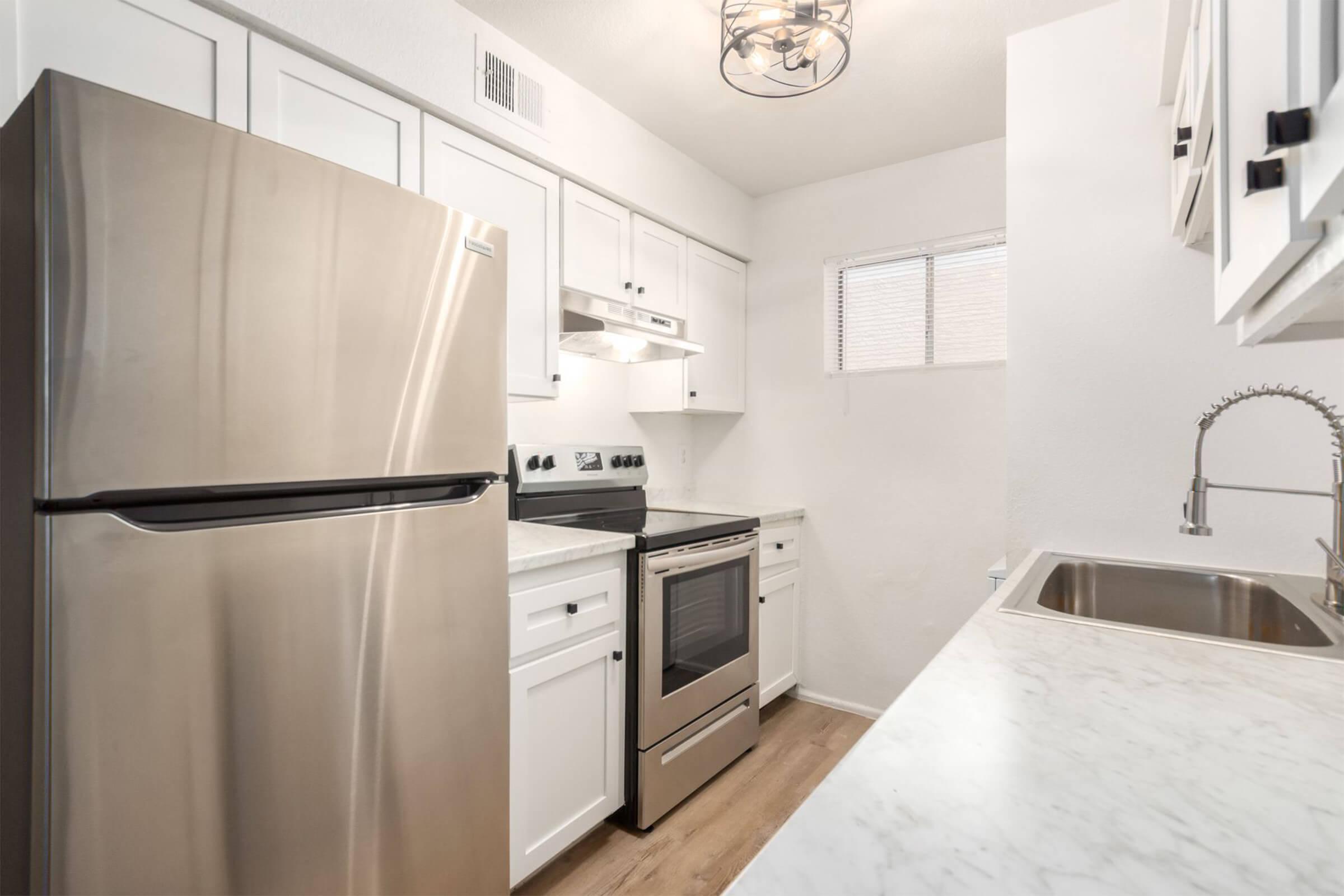 Modern kitchen featuring stainless steel appliances, including a refrigerator, oven, and dishwasher. The white cabinetry contrasts with a marble countertop. A window provides natural light, and a stylish light fixture hangs from the ceiling, enhancing the contemporary decor.