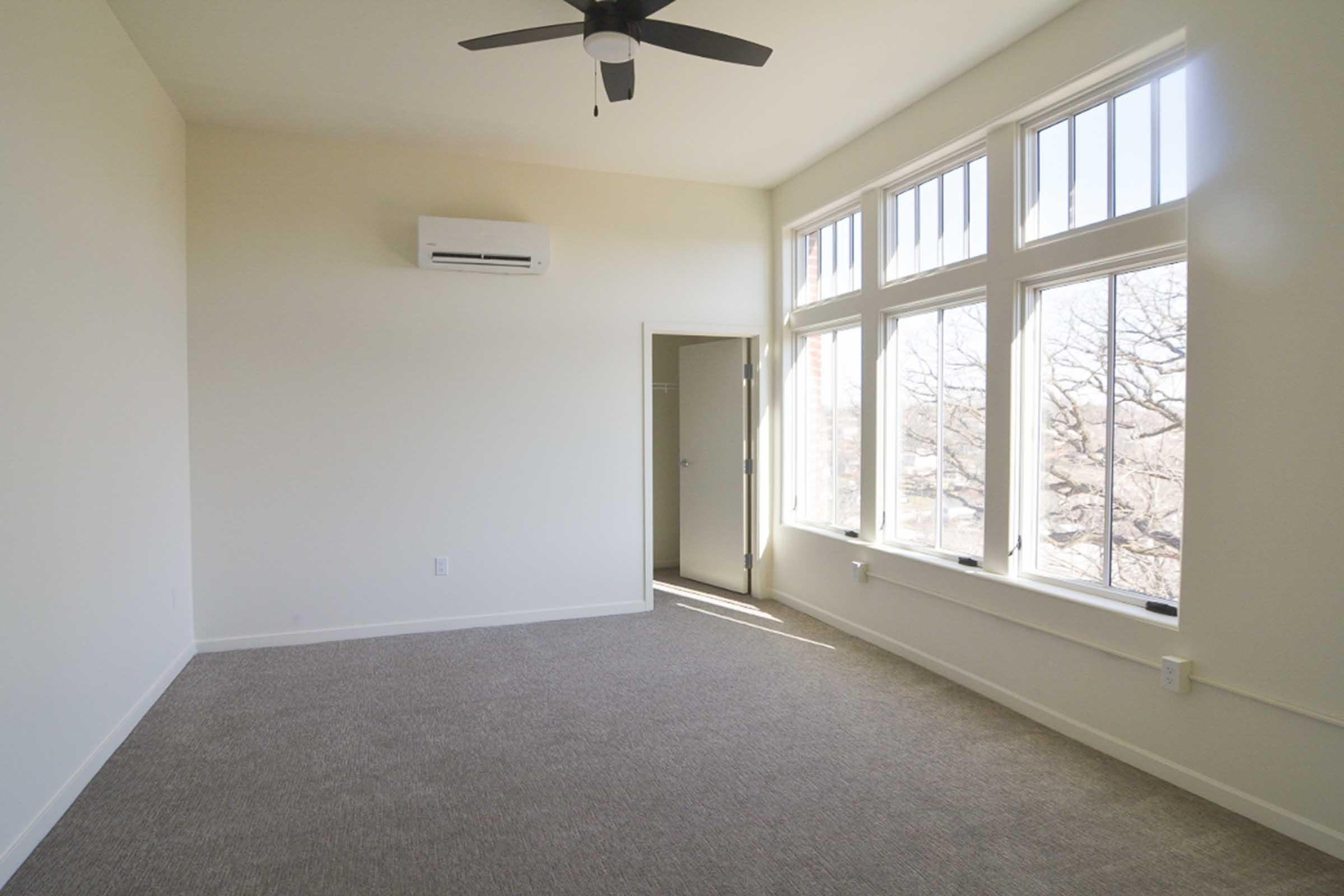 A spacious, empty room featuring large windows that let in natural light, a ceiling fan, and beige carpet flooring. A door on the left leads to another space, while an air conditioning unit is mounted on the wall. The walls are painted a soft white, creating a bright and airy atmosphere.