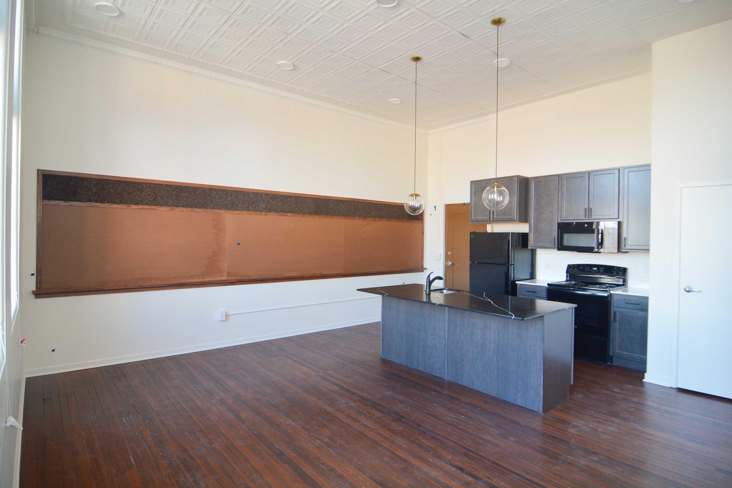 Modern kitchen and living area with a polished wooden floor. Features include a central island with dark cabinetry, stainless steel appliances, and an expansive chalkboard wall. Ample natural light from large windows enhances the open layout.