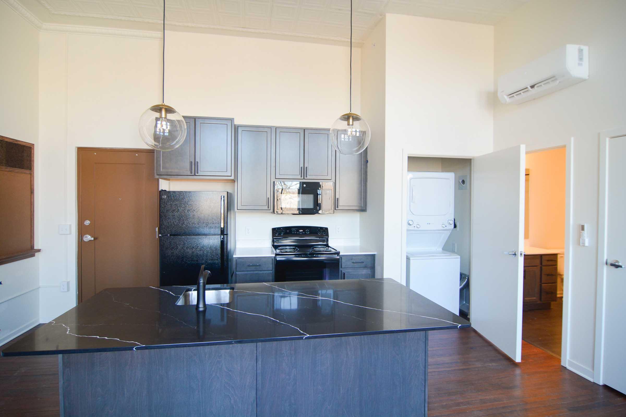 Modern kitchen featuring a large black island with a sink, pendant lighting, dark cabinetry, a black refrigerator, and an oven. A washer and dryer are visible in a nearby room. The space has light-colored walls and wooden flooring, with an open layout that provides a welcoming atmosphere.