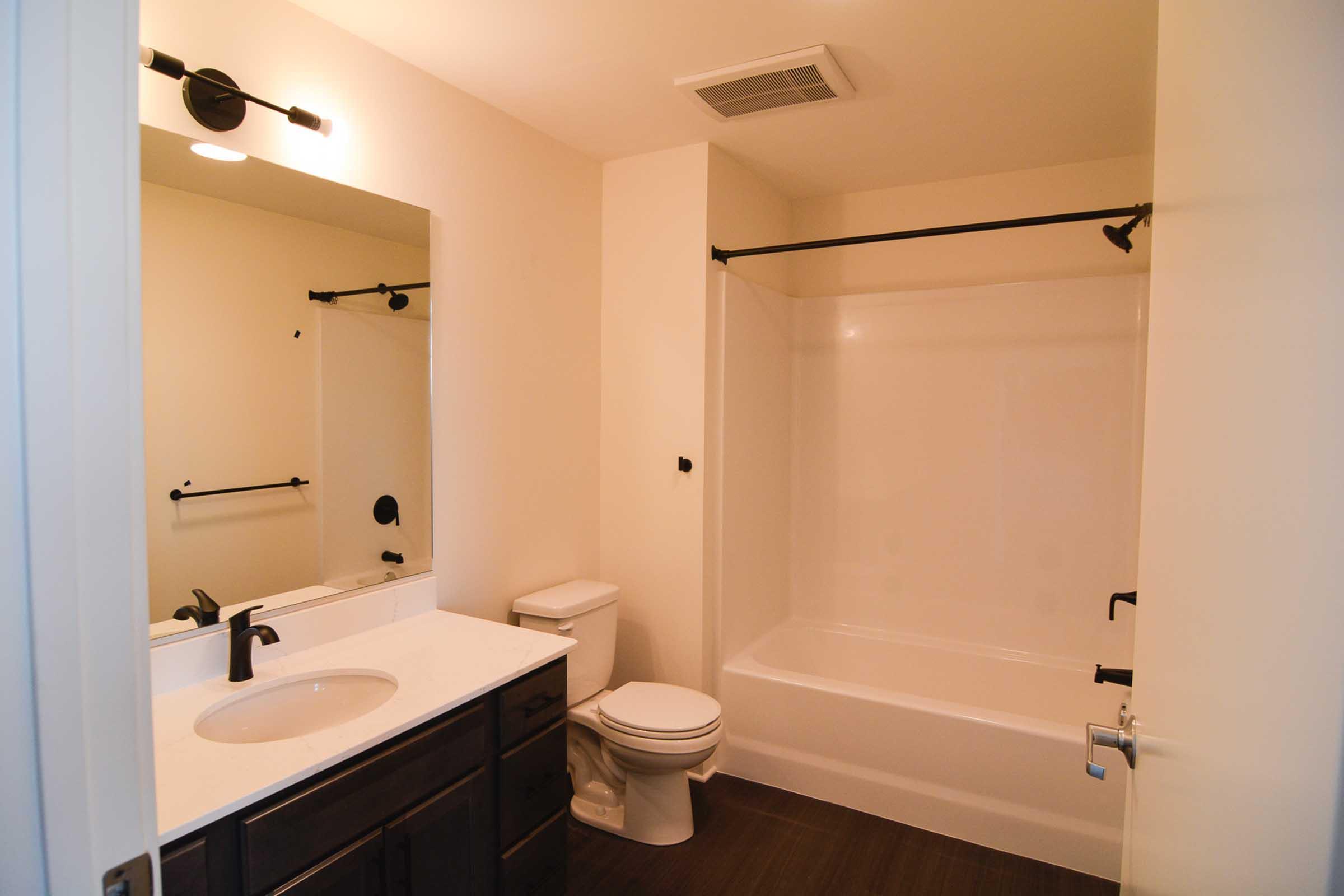 A clean, modern bathroom featuring a white bathtub with a shower curtain rod, a black faucet, a white toilet, and a dark wooden vanity with a sink. The walls are a neutral color, and there is a mirror above the vanity. The flooring is dark, giving a contemporary look.