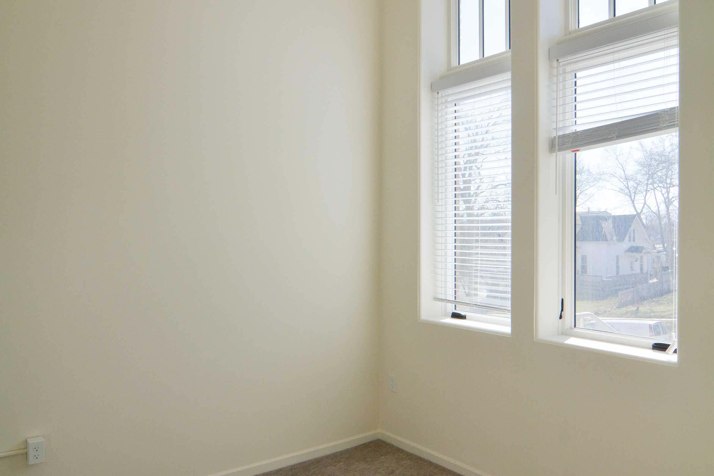 A bright, minimalistic room featuring light-colored walls and large windows with blinds. The carpeted floor is visible, and there's a view of a garden or exterior area outside the windows. The overall ambiance is airy and spacious, ideal for a cozy living space or office.