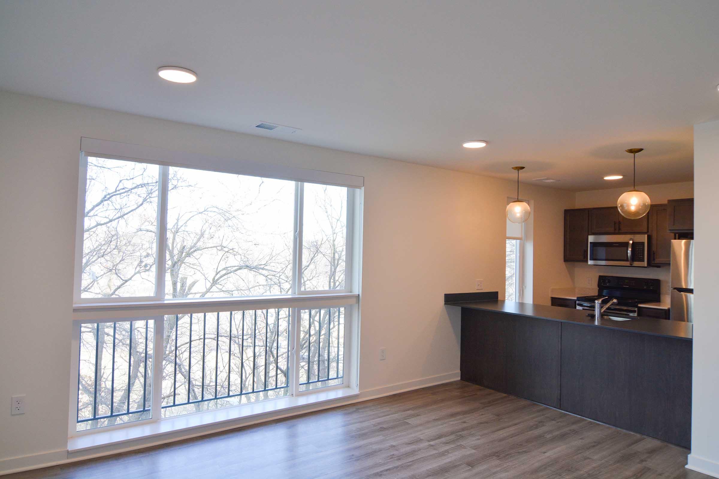 A bright, modern apartment interior featuring a spacious living area with large windows that offer natural light. It includes a kitchen with dark cabinetry and stainless steel appliances, along with pendant lighting. The flooring is light wood, creating a warm and inviting atmosphere.