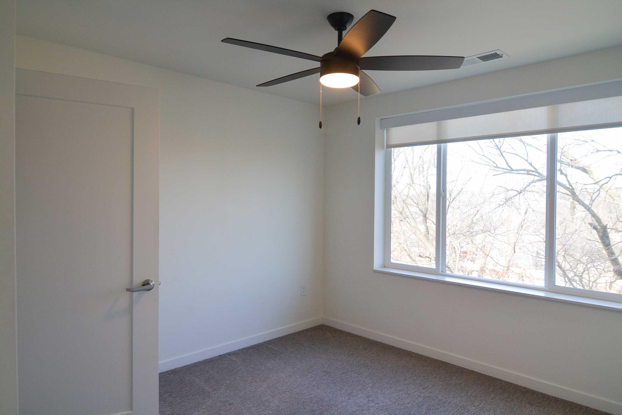 A minimalist bedroom featuring light walls, a ceiling fan with a light fixture, a large window letting in natural light, and neutral-colored carpeting. The room is empty with a simple door visible. Tree branches are visible outside the window, indicating a view of nature.