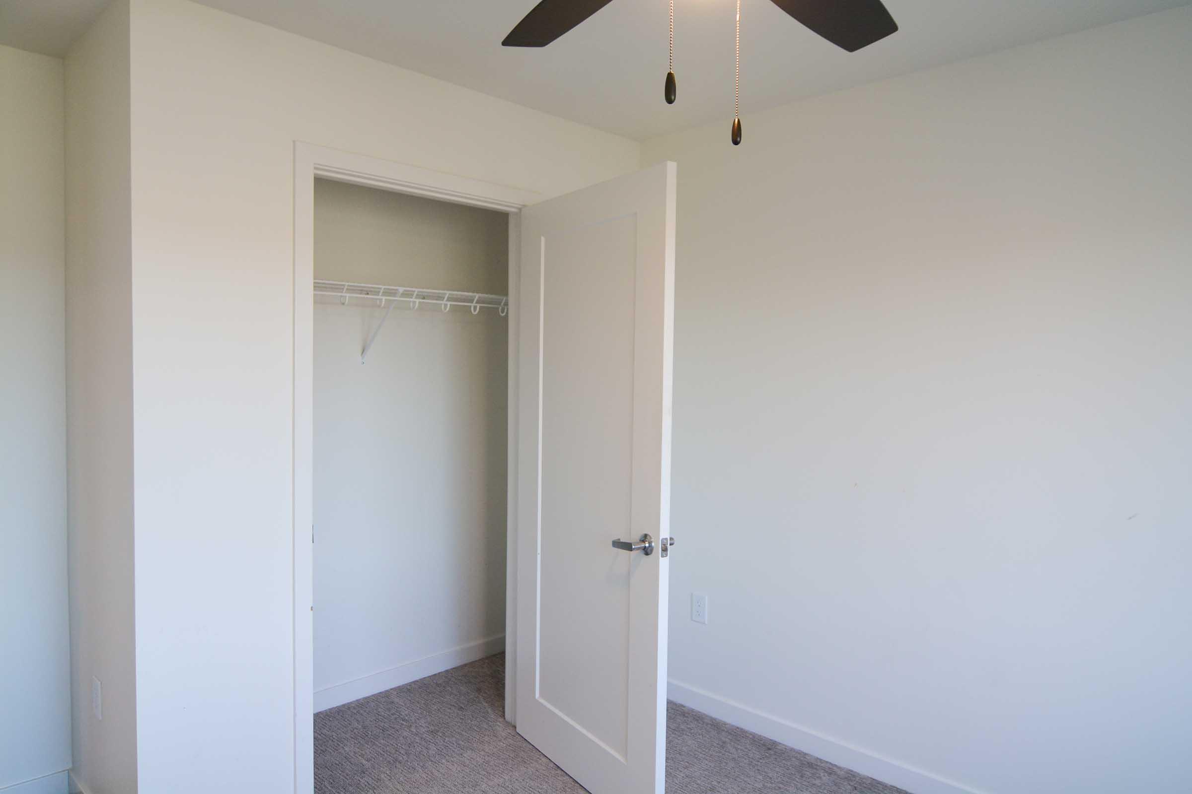 A bright, empty room featuring a white door that is partially open, leading to a closet with a hanging rod. The walls are painted a light color, and the floor is covered in beige carpet. A ceiling fan with black blades hangs above.