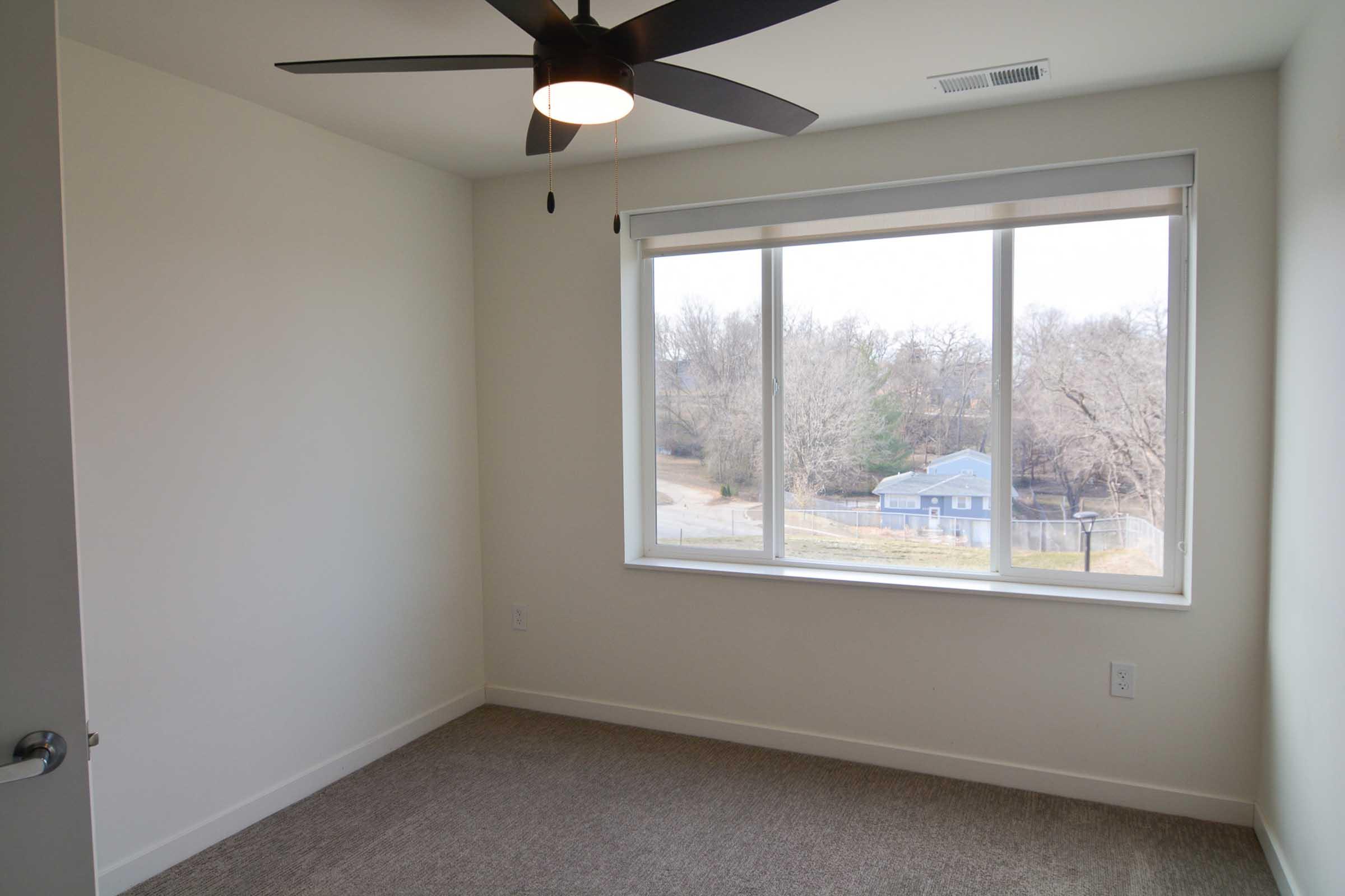 Empty room with light-colored walls and a large window revealing a view of trees and a blue house outside. A ceiling fan is mounted above, and the floor is carpeted in a neutral tone. The overall atmosphere is bright and spacious.
