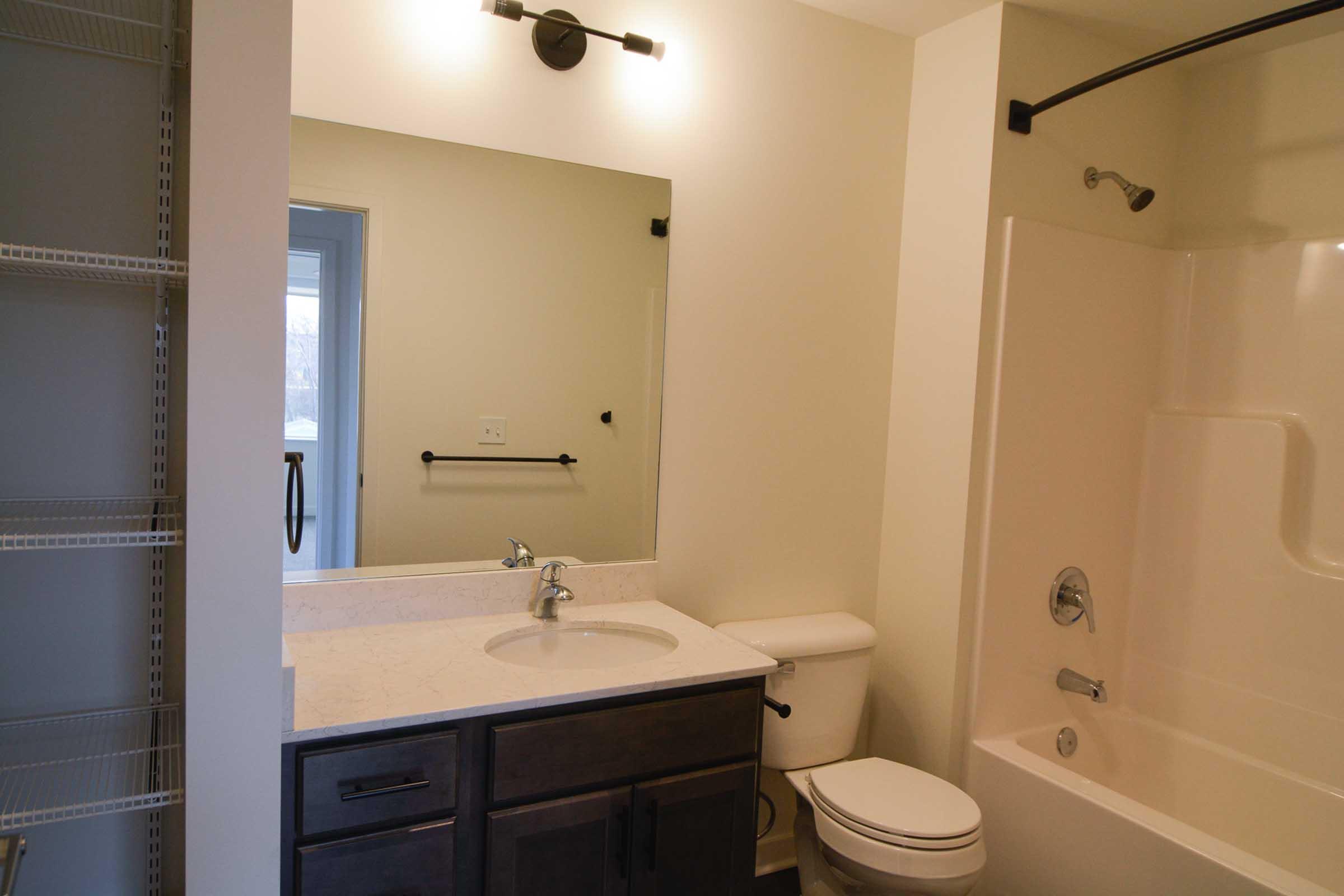 A modern bathroom featuring a white bathtub with a shower, a beige wall, a black towel bar, a large mirror above a sink with a dark wood vanity, and a toilet. There’s a shelf area for storage on the left side of the image. The lighting is warm and inviting, creating a clean and simple aesthetic.