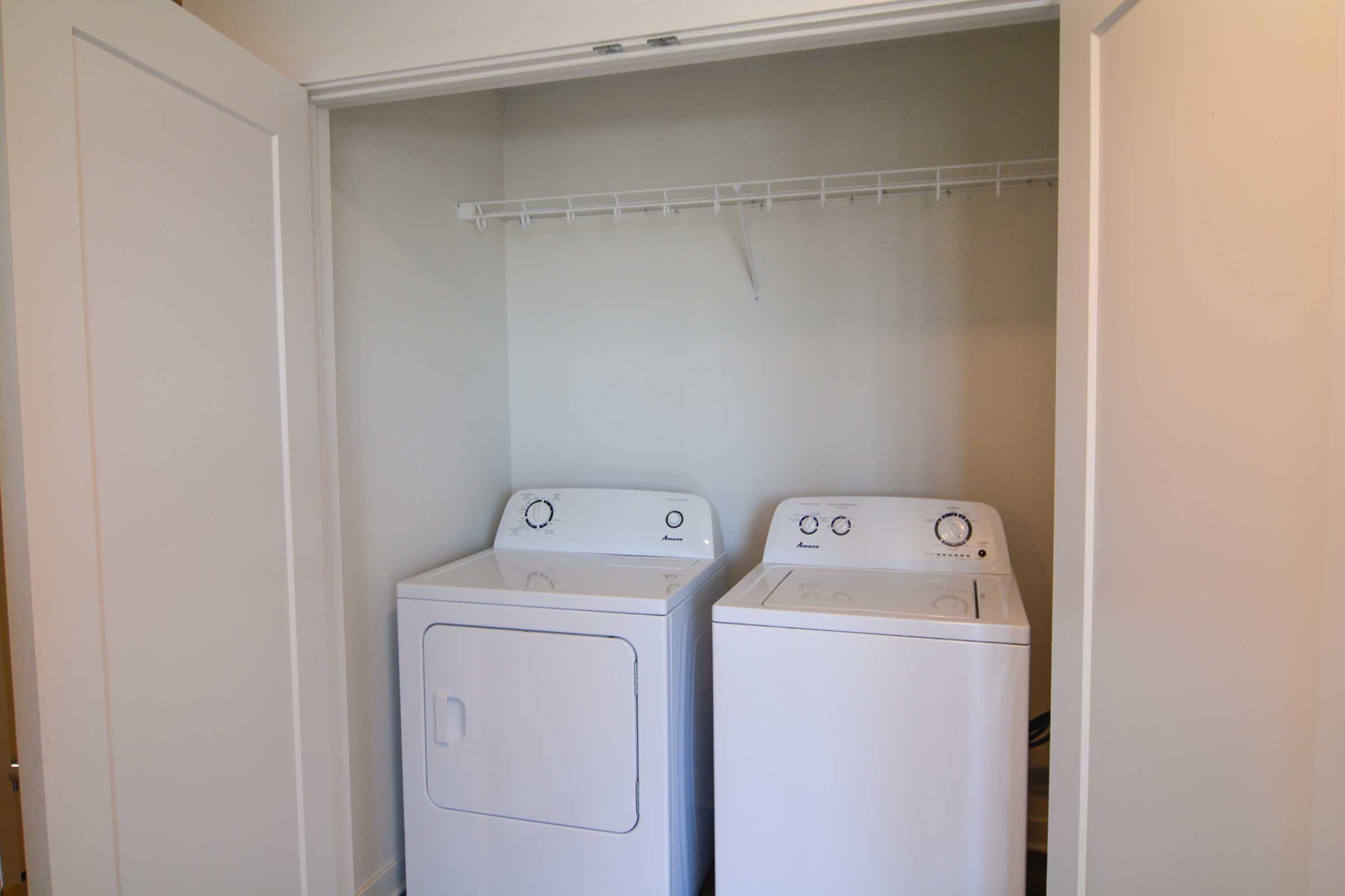 A small laundry area featuring a stackable washer and dryer set. The appliances are white and positioned side by side within a closet space. There is a shelf above them for storage, and the walls are painted a light color, creating a clean and organized look.