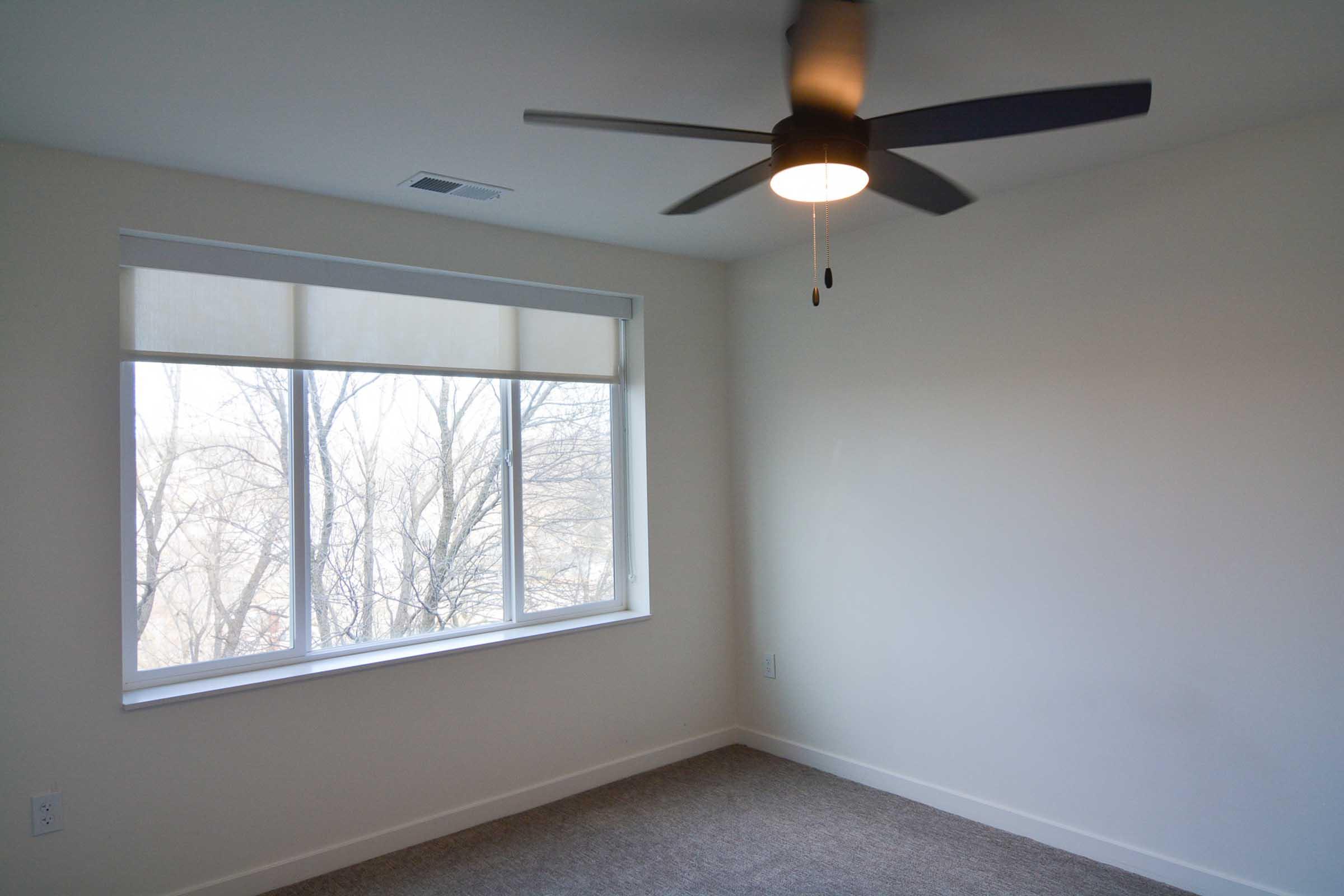Bright, empty room featuring a light-colored wall and carpeted floor. A large window with a white blind allows natural light, revealing trees outside. A modern ceiling fan is installed, enhancing the room's airy feel. The overall aesthetic is minimalist and inviting.