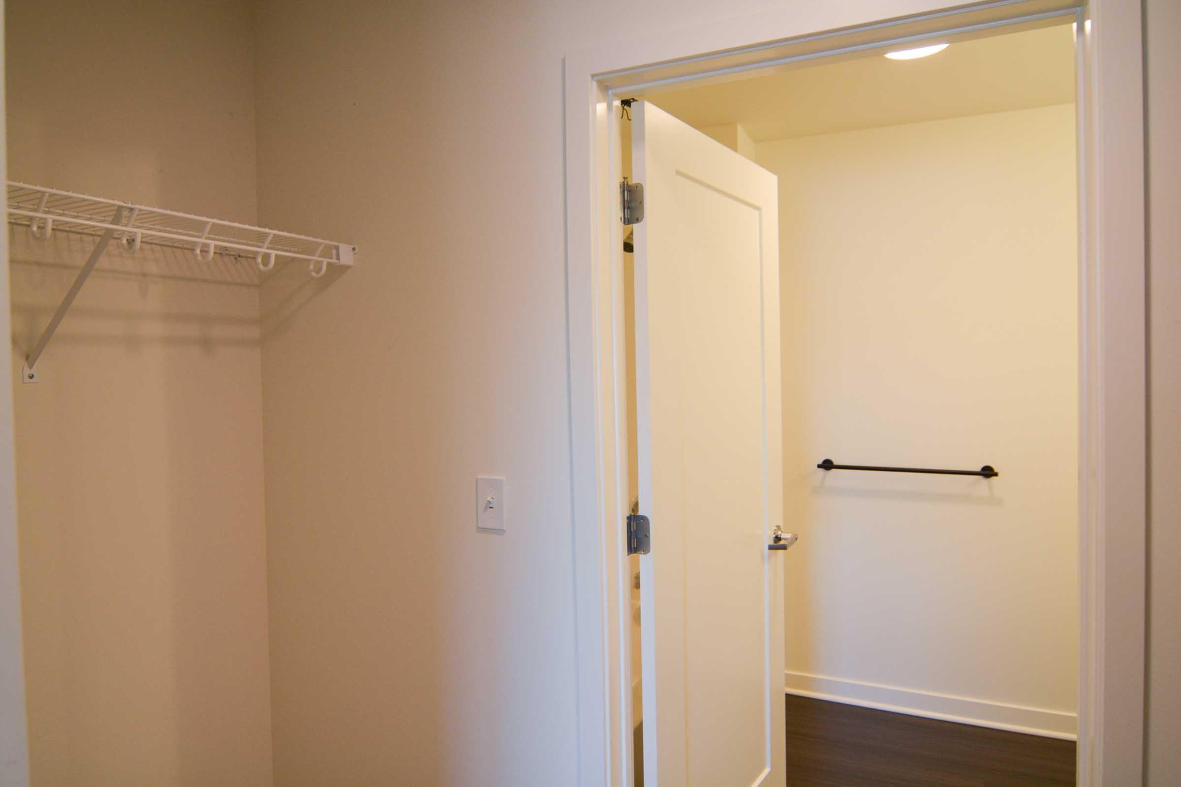 A view of a light-colored room with an open door leading to a small closet. The closet features a clothes rod and a shelf for storage. The interior walls are painted in a neutral tone, and the flooring is dark wood. Natural light illuminates the space, creating a bright and inviting atmosphere.