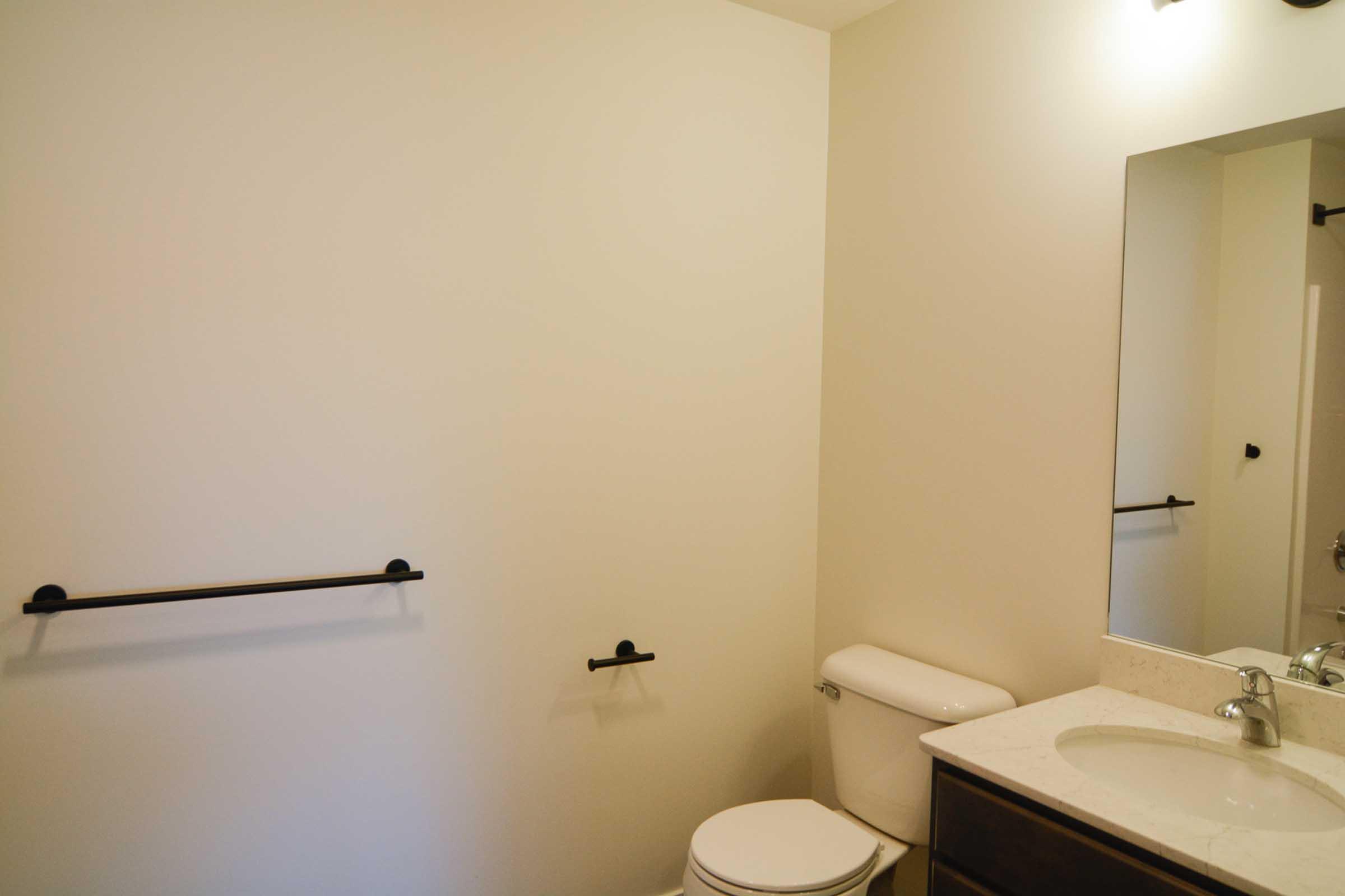 A clean and modern bathroom featuring a white wall, a toilet, a large mirror above a sink, and a towel rack. The countertop is light-colored, and the fixtures are simple and elegant, creating a neutral and spacious atmosphere.