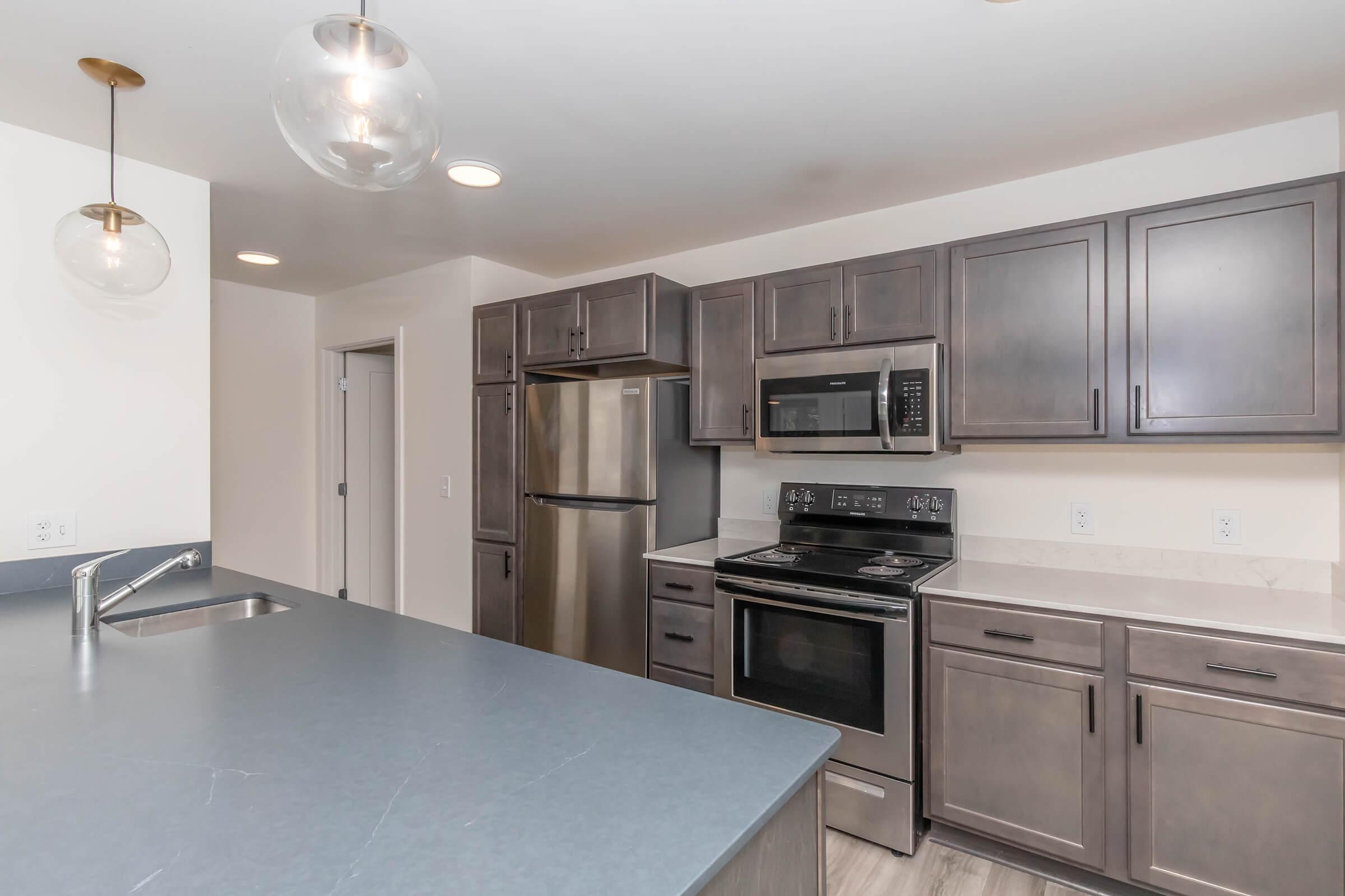 Modern kitchen featuring stainless steel appliances, including a refrigerator, microwave, and stove. Dark cabinetry contrasts with light countertops. An island with a sink provides additional workspace, and pendant lighting adds a stylish touch. The open layout is bright and inviting.