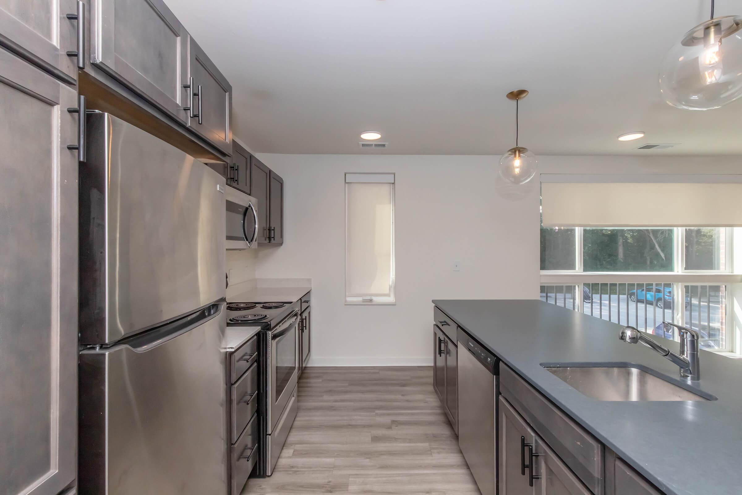 Modern kitchen featuring stainless steel appliances, including a refrigerator, oven, and microwave. A sleek countertop with a single basin sink and faucet runs along one side. The space is illuminated by pendant lighting, with large windows letting in natural light. Light-colored cabinetry complements the overall design.