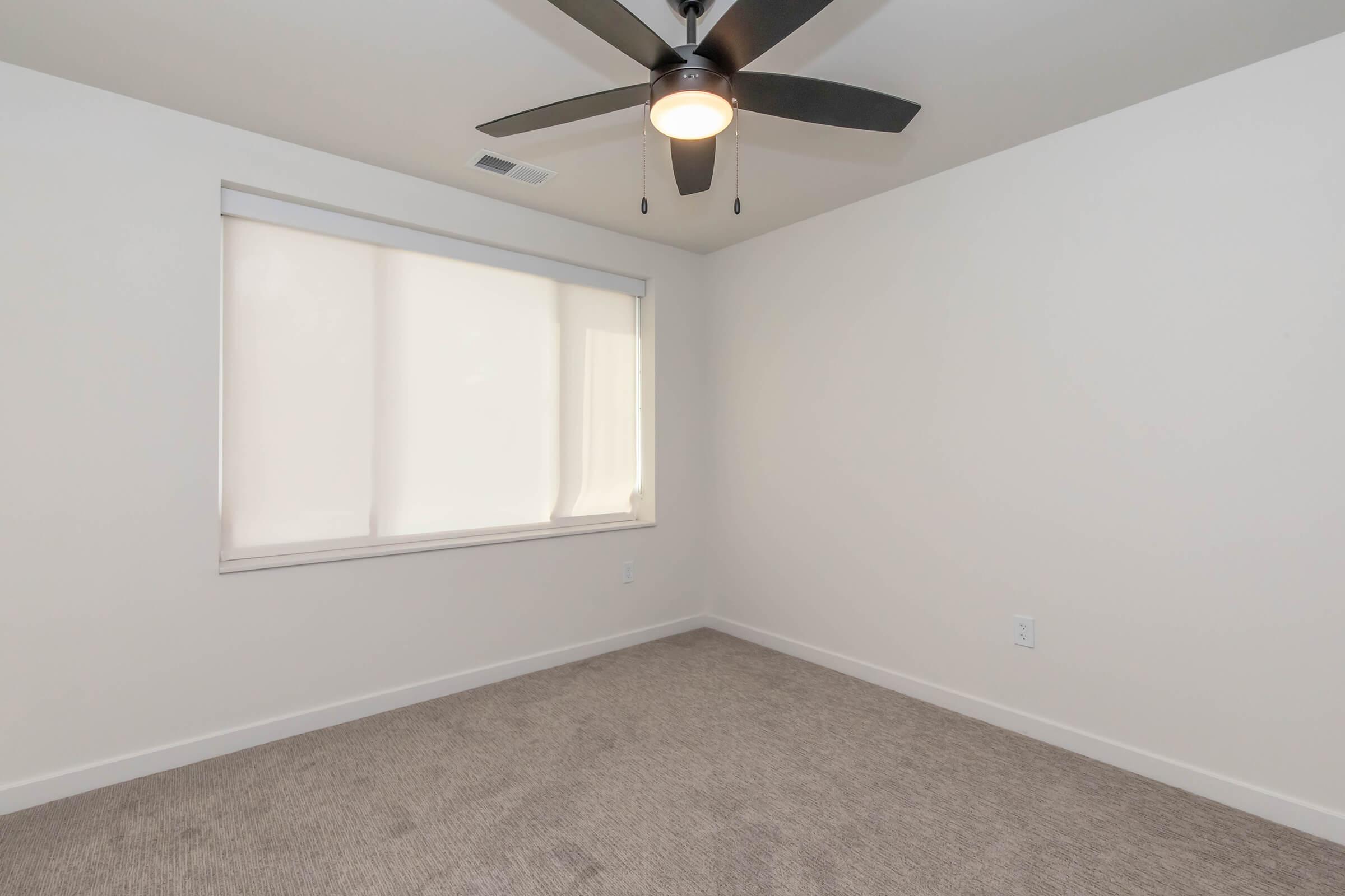 A small, empty room with light beige carpet and white walls. A ceiling fan with a light fixture is mounted on the ceiling. A window covered with a sheer white shade allows natural light to enter, creating a bright and airy atmosphere. The space is unfurnished, emphasizing its simplicity.