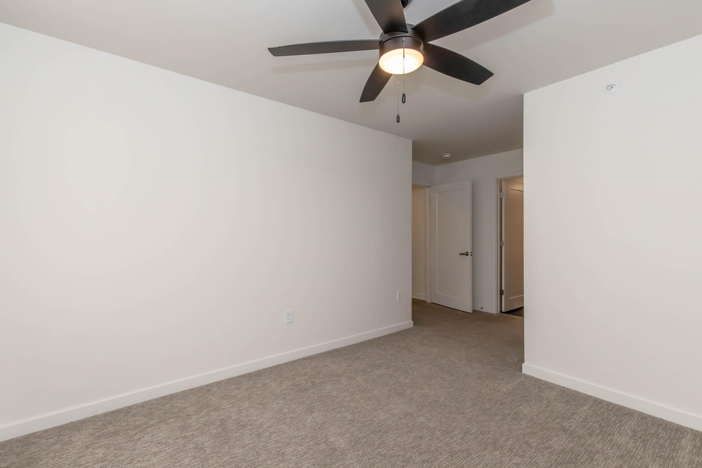Empty room with light-colored walls and a ceiling fan. The floor is carpeted in a neutral tone. A doorway is visible on the right, leading to another room. The space is well-lit and appears modern and minimalist.