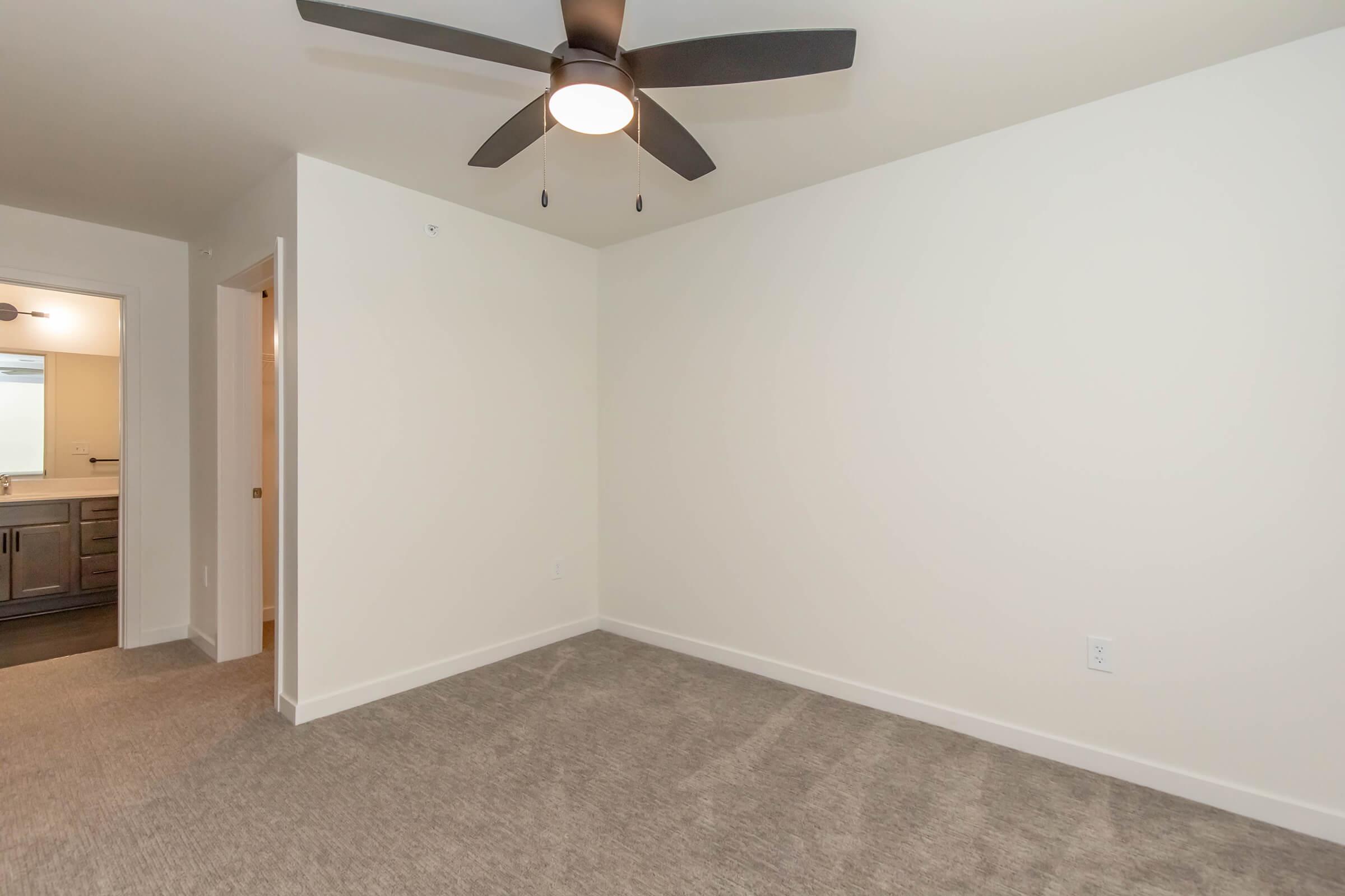 A spacious, empty room with light-colored walls and a ceiling fan. The floor is covered with gray carpeting, and there’s an open doorway leading to another area. The room has a clean, modern look, suitable for various uses.