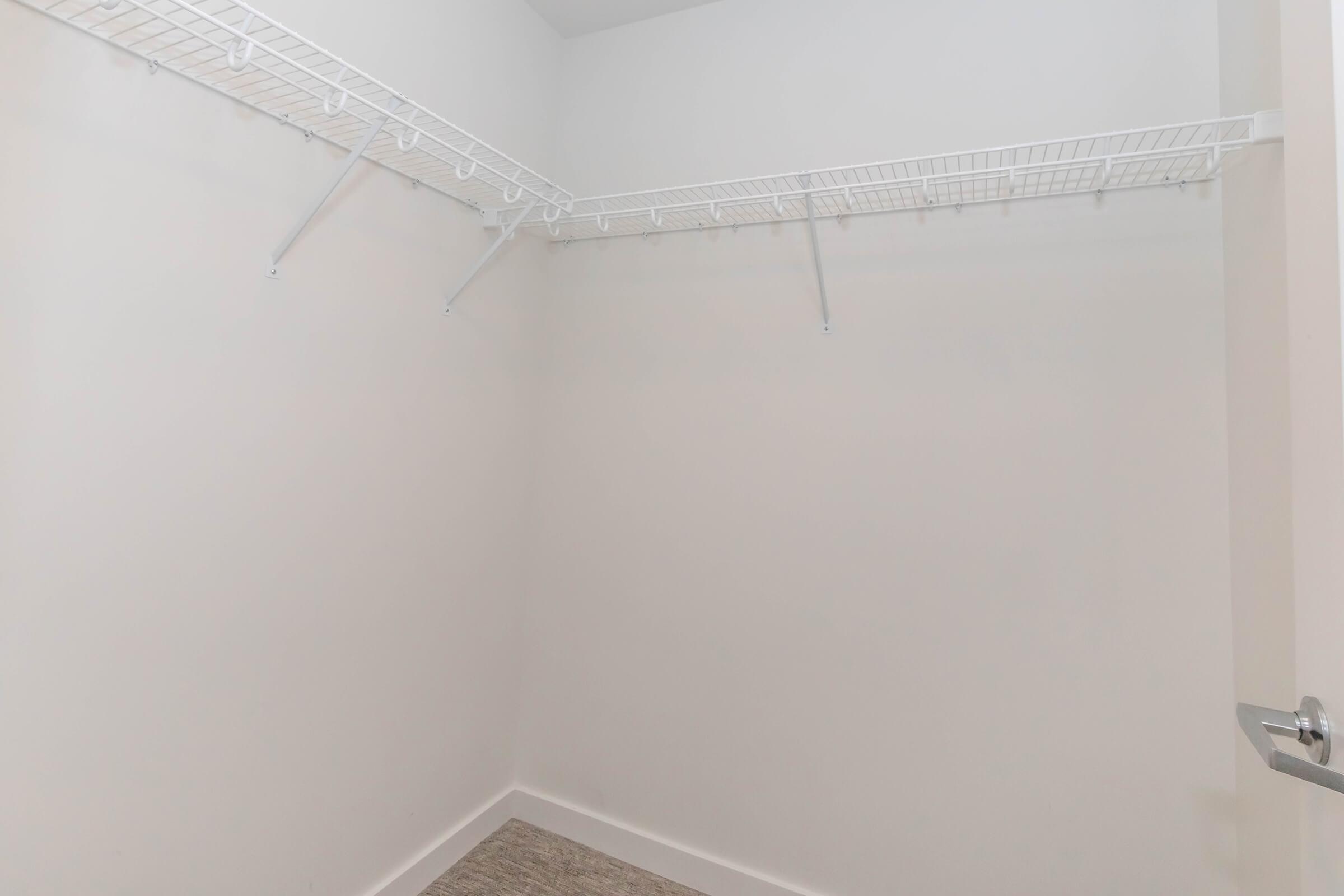 Empty closet with white walls, a light gray floor, and a wire shelving unit along the back wall. There are no items or clothing hanging on the shelves, creating a spacious and minimalistic appearance. A partially open door is visible on the right side.