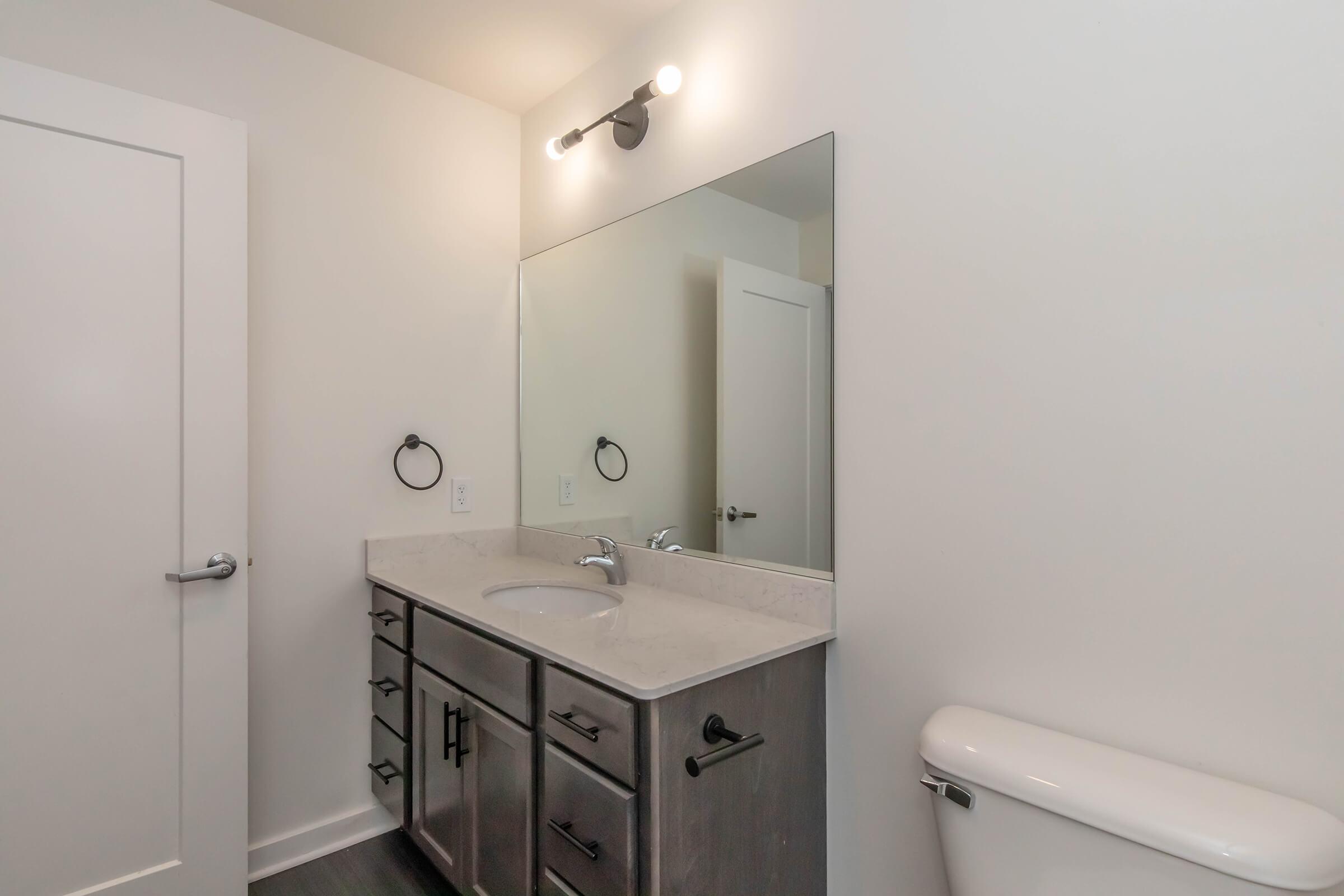 A modern bathroom featuring a sleek vanity with a white countertop and dark wood cabinets. There is a large mirror above the sink, complemented by stylish wall-mounted lighting. A white toilet is positioned nearby, and the walls are painted in a light color, creating a clean and spacious look.