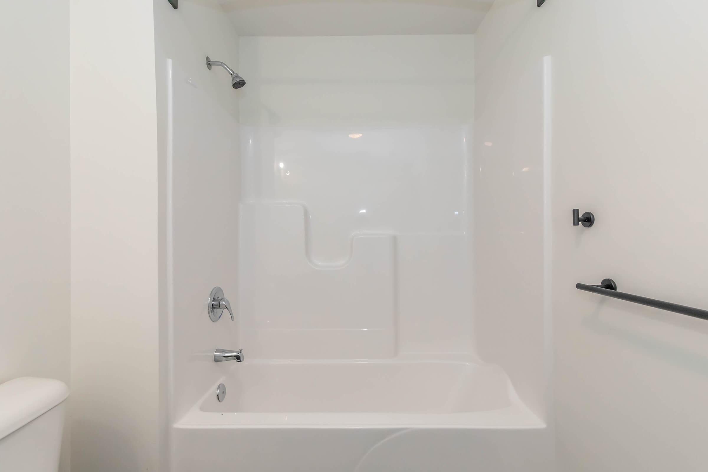 A clean, modern bathtub and shower combination with white acrylic walls. The shower features a handheld showerhead and a wall-mounted faucet. A safety grab bar is installed beside the tub for support. The overall design is minimalistic and functional, set against a light-colored wall.