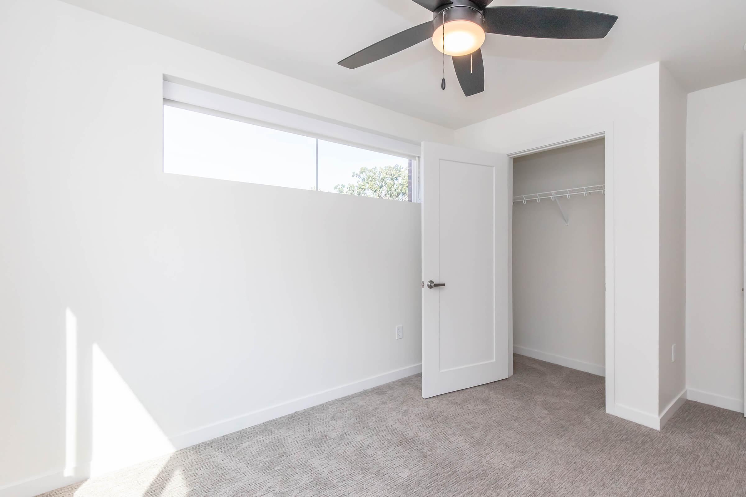 A bright, minimalist bedroom with light gray walls and plush carpet. It features a ceiling fan and a large window allowing natural light to enter. There is a door leading to a closet with a hanging rod, and the room has an open and airy feel, ideal for relaxation and comfort.